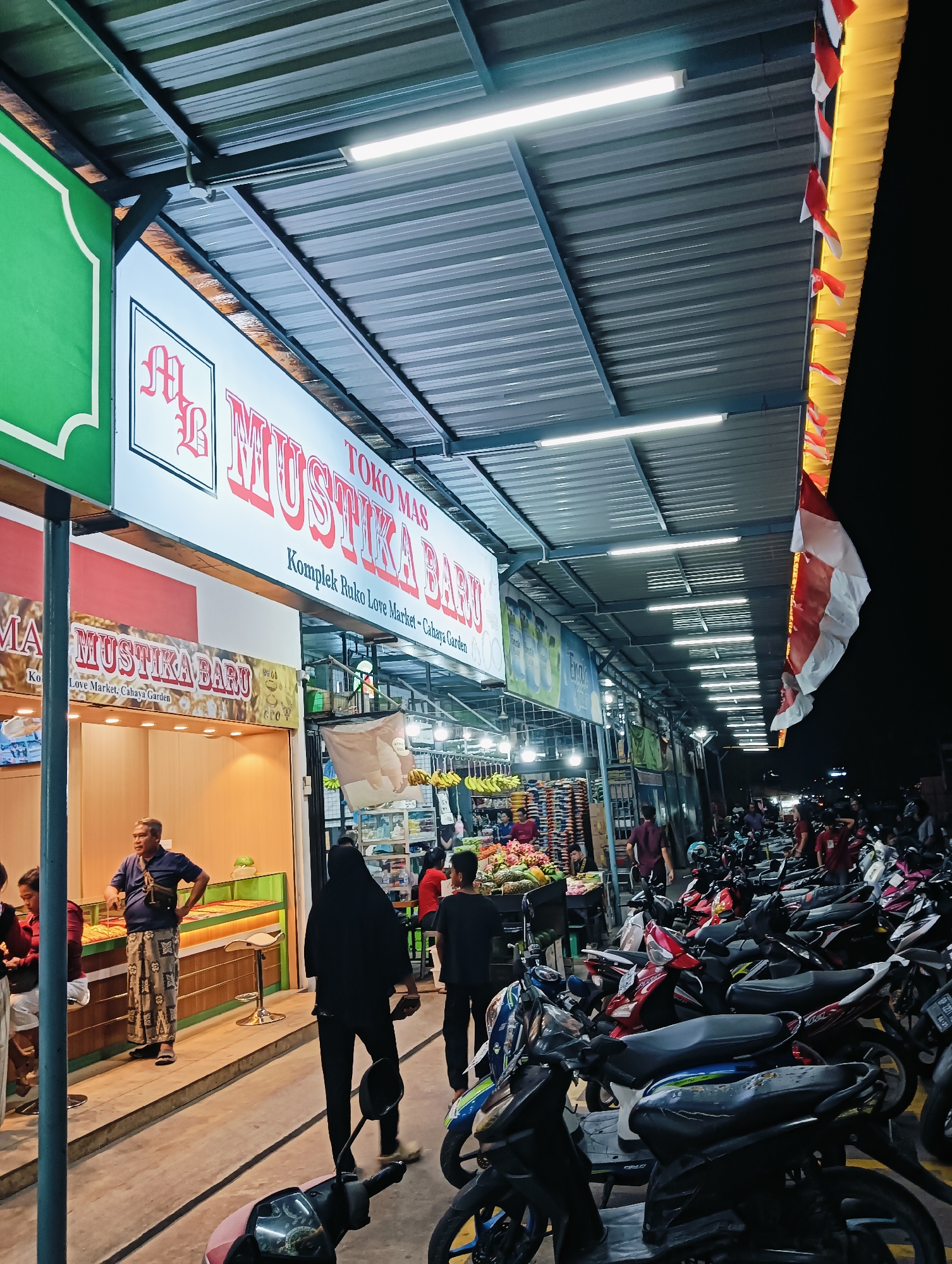 The image depicts an outdoor market scene at night in Batam, Indonesia, specifically in front of "Toko Mas Mustika Baru" at Komplek Ruko Love Market - Cahaya Garden. The foreground is dominated by parked motorcycles, with several individuals walking or standing around. A prominent sign for "Toko Mas Mustika Baru" hangs above a stall displaying goods. Further down the market, there are stalls with produce, including fruits and what appear to be stacked goods. The market is illuminated by overhead fluorescent lights and decorative yellow lighting with small Indonesian flags. The overall atmosphere suggests a bustling night market or commercial area.