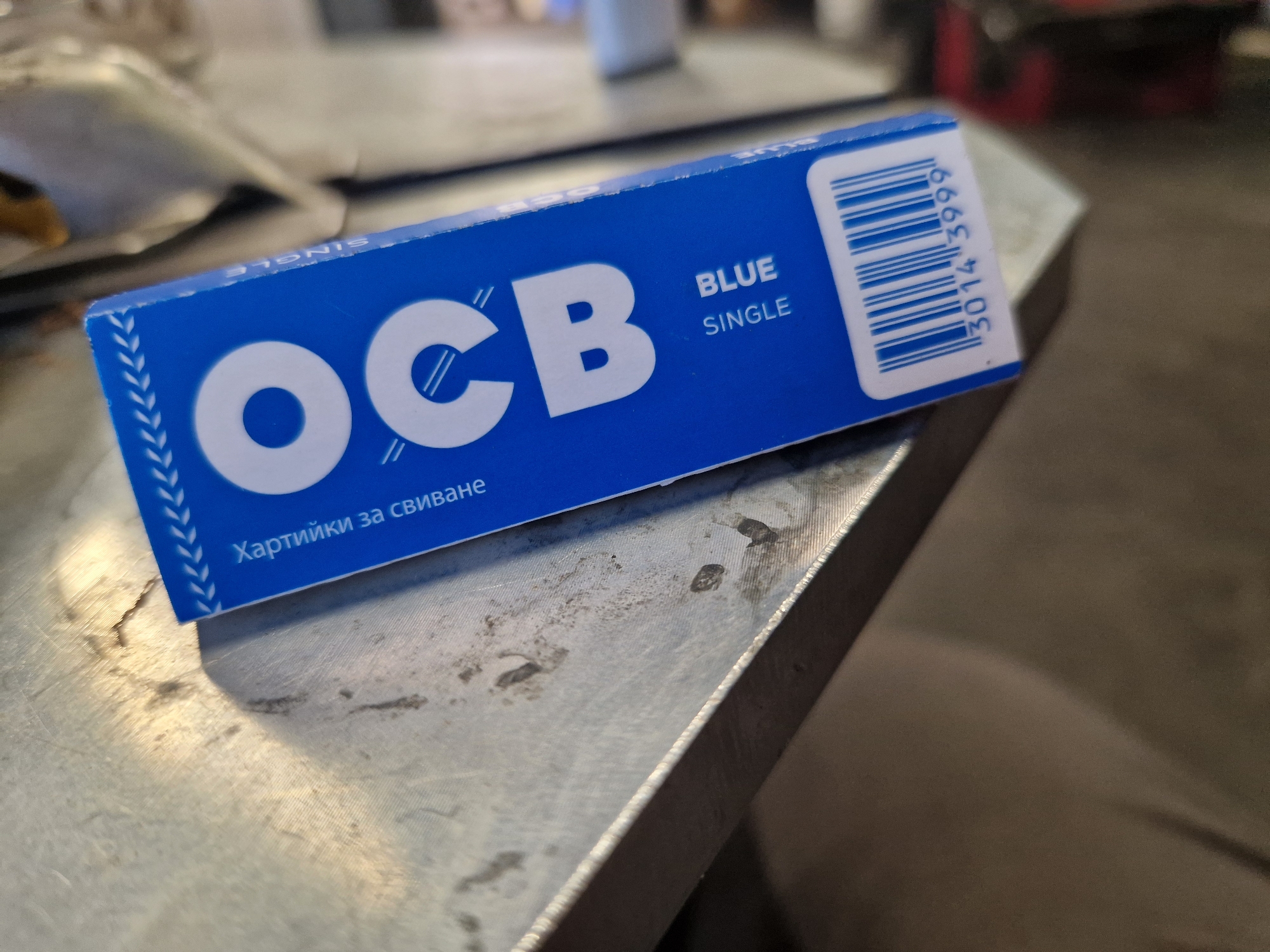 The media file presents a close-up view of a blue packet of OCB rolling papers, resting on a textured, metallic surface. The surface appears somewhat worn, with visible scratches and smudges, suggesting a utilitarian environment like a workbench or industrial table. The background is out of focus, showing blurred shapes and colors, consistent with an indoor setting.
The packet itself is bright blue with clear white text. The brand name "OCB" is prominently displayed in large capital letters. Below and to the right of the brand, the words "BLUE" and "SINGLE" are printed, describing the product variant. Along the lower edge of the packet, the Bulgarian text "Хартийки за свиване" is visible, which translates to "Rolling papers," providing a specific location cue consistent with Sofia, Bulgaria. A white barcode with the numbers "30143999" is partially visible on the right side of the packet. No individuals or specific activities are shown; the image is a static product shot under artificial indoor lighting.