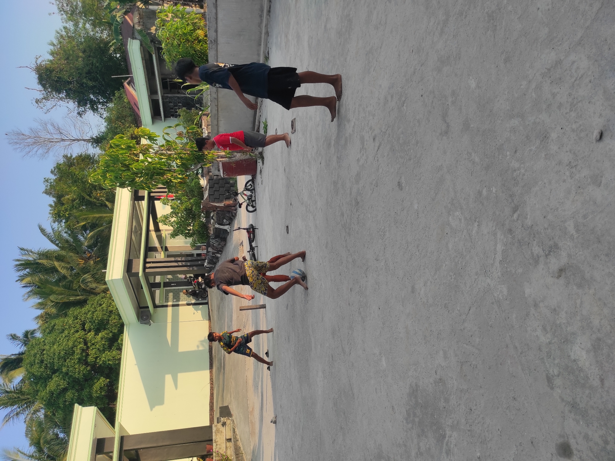 In an outdoor, open-air, concrete-paved area, likely a yard or courtyard, four young males are engaged in a game of soccer. The setting appears to be a residential or possibly a small commercial area, with buildings and lush green trees visible in the background. The architecture suggests a warm climate, possibly in a tropical or subtropical region. The sky is clear and blue, indicating daytime.
Two of the individuals are actively playing with a soccer ball. One, wearing a dark t-shirt and yellow patterned shorts, is positioned to the right of the ball, appearing to kick it. Another, in a black and yellow patterned shirt with the number 56 on his shorts, is on the left, also seemingly involved in the play.
Two other individuals are observing or waiting for their turn. One, on the far right, wears a dark t-shirt and black shorts, standing with his back to the camera. To the left of him, a younger boy in a red t-shirt and grey shorts stands watching.
Scattered on the ground are a few objects, including a small, red barrel or container, and a bicycle. The concrete surface shows some wear and tear, with faint marks and a few small stones. The overall scene is one of casual recreation in a bright, outdoor environment.