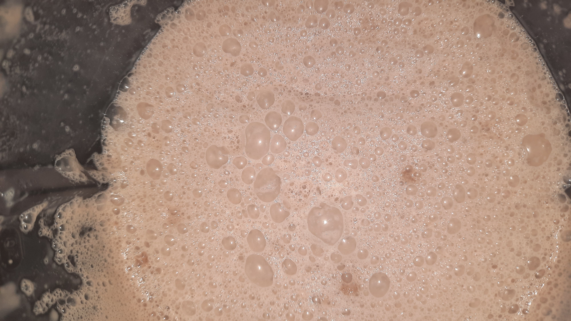 The image shows a close-up view of a frothy, light brown liquid in a dark container. The surface is covered with numerous bubbles of varying sizes, some appearing larger and rounder, while others are smaller and more irregular. The liquid appears to be thick and possibly creamy. The top edge of a dark, possibly plastic or metal, container is visible along the upper part of the image. The background is dark and out of focus. There is no indication of a city or any specific location within Шклоў, Belarus. The image is a still photograph with no discernible action or interaction.