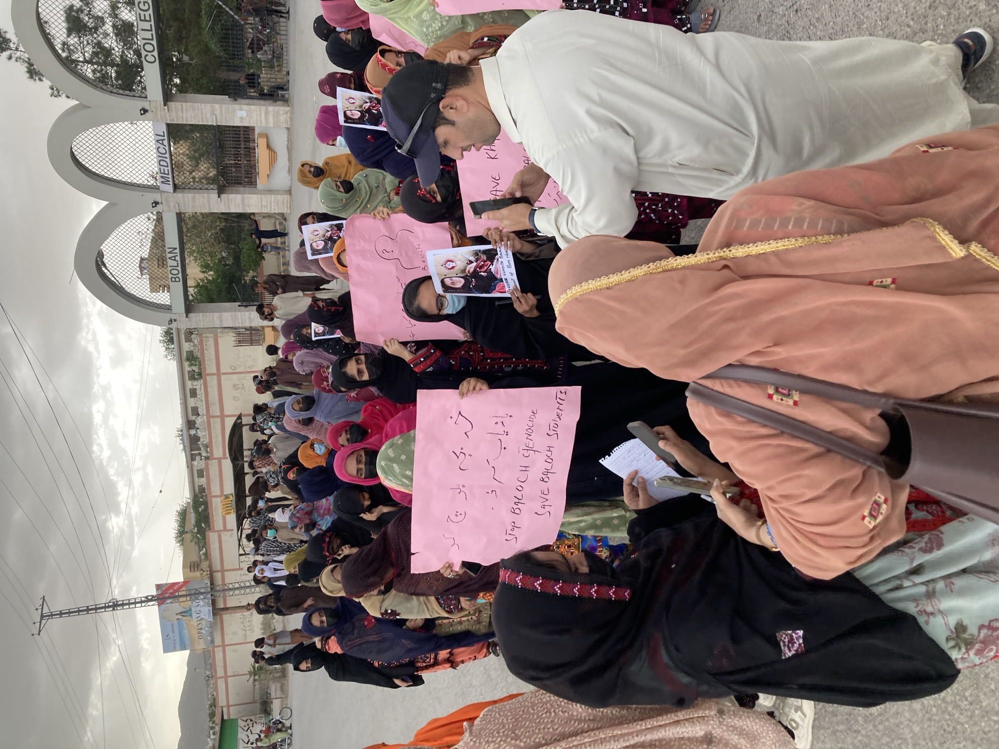 A large group of people, primarily women, are seated on the ground outdoors, participating in a protest or demonstration. Many are wearing traditional clothing including headscarves and face masks. One man, wearing a white shalwar kameez and a black cap, is visible in the foreground, holding a mobile phone.
The event is taking place on a paved area in front of an institutional building. Large arched signs overhead clearly identify the location as "BOLAN MEDICAL COLLEGE". In the background, there are trees, a fence, and distant mountains under an overcast sky. A billboard further in the distance advertises "OPENING SOON".
Participants are holding numerous pink placards with handwritten messages. Visible English text on the placards includes "STOP BALOCH GENOCIDE" and "SAVE BALOCH STUDENTS." Other text includes "DELERSE KHADITH BALACH" and "OF BIN NUR". Some placards also feature photographs of individuals. Text in what appears to be Urdu or Balochi script is also present, reading "خہ بچہ بلوچ کو باز یاب کرو۔" which translates to "Find the Baloch child." or "Recover the Baloch child." The collective mood appears serious, indicative of a protest. The scene is set during daytime under cloudy weather conditions, in Quetta City Tehsil, Pakistan.