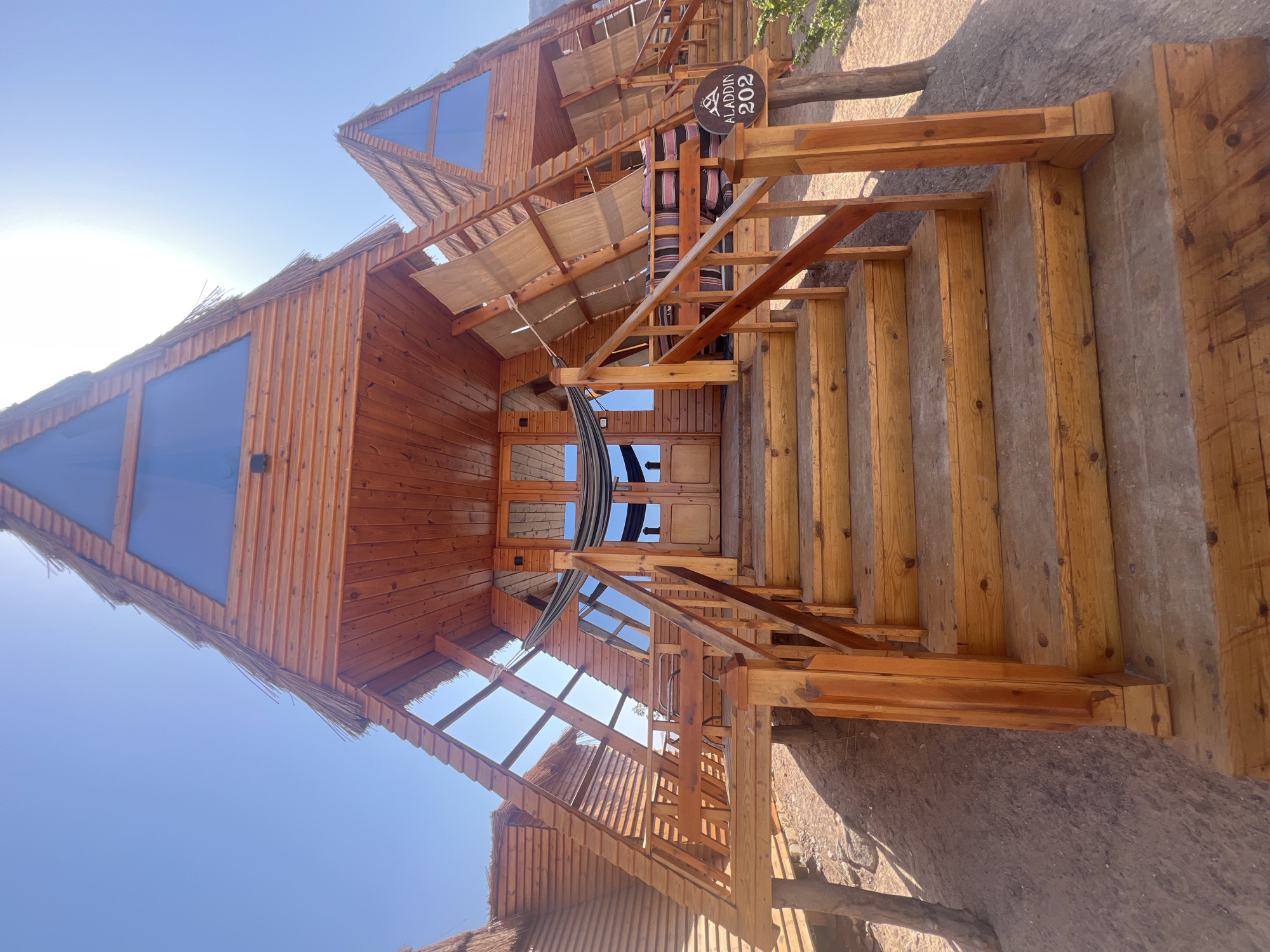 A close-up, eye-level, vertical shot captures the entrance of a wooden, A-frame cabin with a thatched roof, likely in Nuweiba, Egypt. The cabin is elevated on wooden stilts, with a wooden staircase leading up to the main entrance. The stairs are made of rough-hewn wood, with a wooden handrail on the right. At the top of the stairs, a wooden porch features double doors and a hammock strung between two poles. To the right of the entrance, a sign reads "ALADDIN 202." Behind the porch, more A-frame structures are visible, also with thatched roofs and large triangular windows. The scene is bathed in bright sunlight under a clear blue sky. The ground surrounding the cabin is sandy and rocky, typical of a desert or coastal environment. The overall impression is rustic and natural.