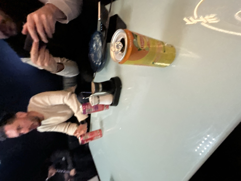 The image depicts an indoor scene at what appears to be a restaurant or bar in Illkirch-Graffenstaden, France, focused on a light-colored, reflective table surface. Three individuals are partially visible in the low-lit background.
On the left, a male subject with dark hair and a beard, wearing a light-colored long-sleeve shirt, is seated, with his mouth slightly open, suggesting he is speaking or reacting. Two red or pink beverage cans, one appearing to be a Red Bull, are situated near his position on the table.
On the right, another male subject, wearing a dark vest over a light long-sleeve shirt, is seen holding a dark rectangular object, likely a smartphone, with both hands, indicating interaction with the device.
In the immediate foreground, on the right side of the table, a yellow Lipton Iced Tea (Peach flavor) can, with an open tab, stands upright. Further back on the table, centrally located, is a black caddy holding white salt and dark pepper shakers. A dark blue or patterned plate or bowl, and a dark rectangular object (possibly another phone or menu) are also visible on the table. A metal utensil handle is partially visible near the plate. A third individual is vaguely discernible in the far left background, also seated. The environment features dark upholstered seating and artificial lighting, creating reflections on the table surface.