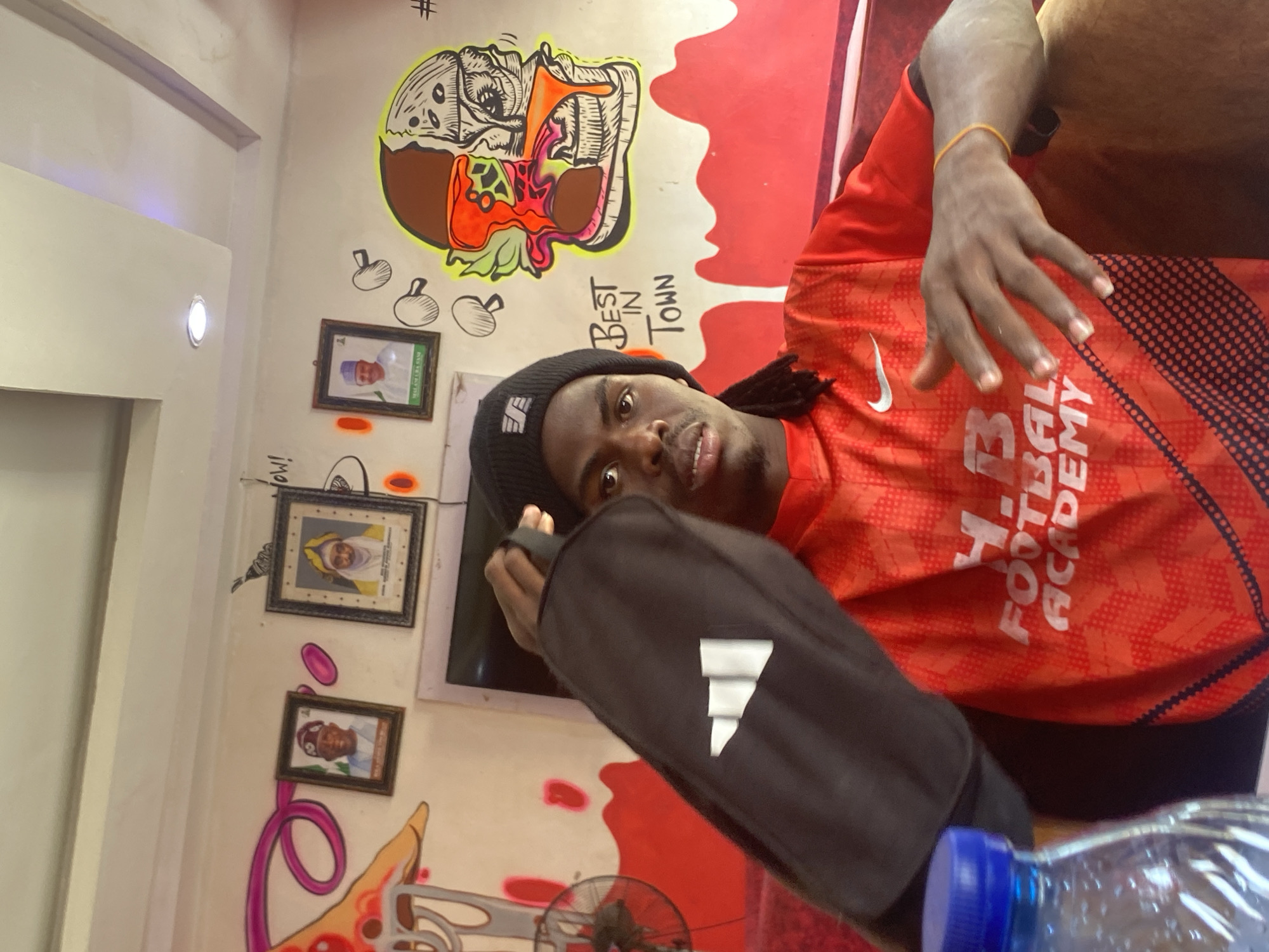 A man is indoors, potentially in a restaurant in Tukur Tukur, Nigeria. He wears a black beanie and an orange athletic jersey. He holds a black Adidas bag. Behind him, the wall is decorated with a cartoon burger, framed portraits, and graffiti. A water bottle sits on the table in the foreground.