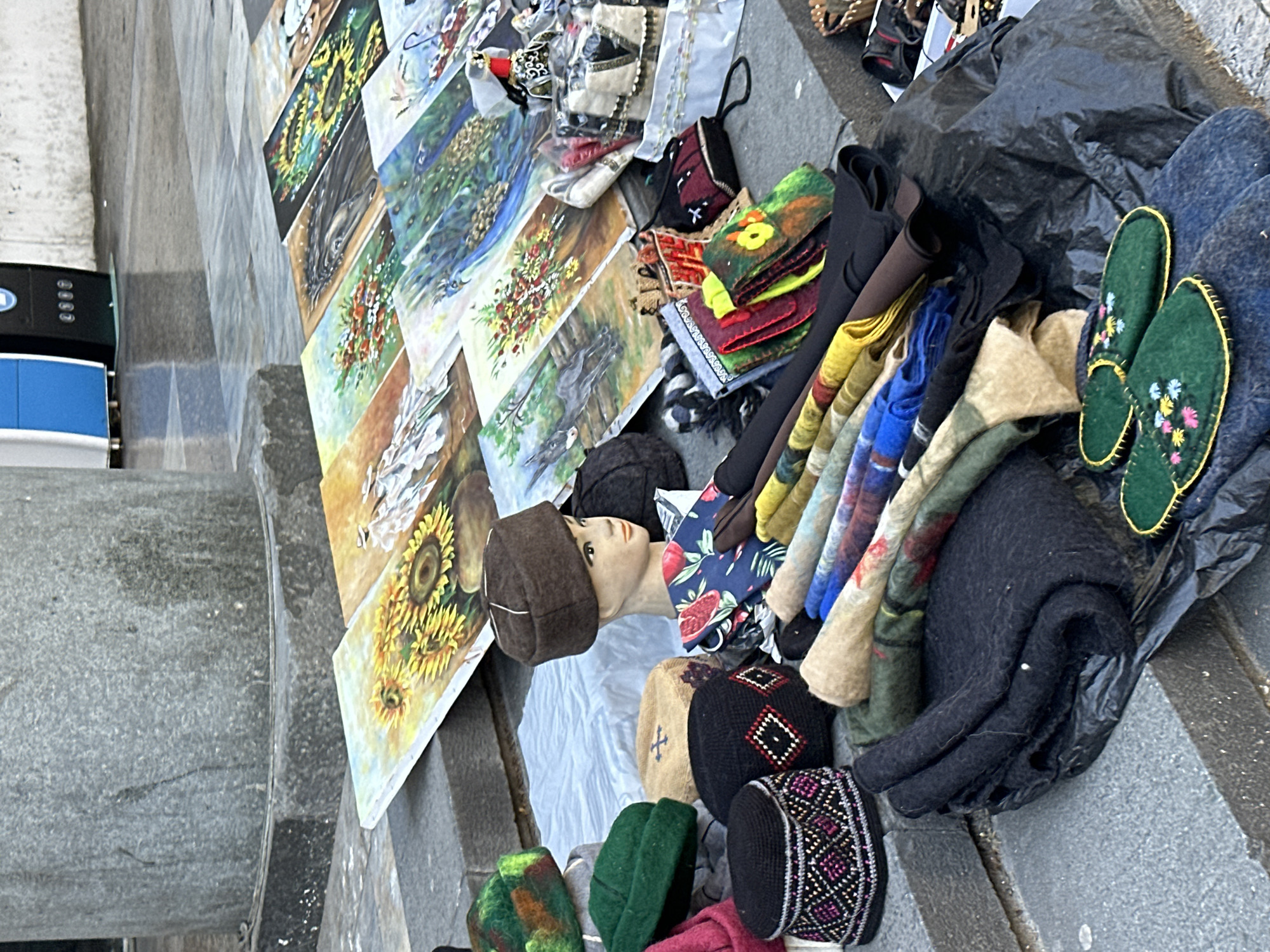 The image depicts a street vendor's display in Tbilisi, Georgia, featuring an assortment of handmade crafts and souvenirs. The items are arranged on a gray stone surface, likely a sidewalk or steps.
The array of goods includes:
* A series of rectangular paintings, leaning against a wall or object to the left, depicting various subjects like sunflowers, flowers, and possibly animals.
* Handmade wool hats and caps of various colors and patterns.
* A mannequin head wearing a brown hat, used to display the hats.
* Colorful scarves or fabric pieces folded and stacked.
* Embroidered green slippers.
* Other miscellaneous items, including possible bags and accessories.
The scene suggests a typical street market atmosphere common in tourist areas, with the vendor presenting their wares for sale. The presence of the stone surface indicates a historic or traditional setting within Tbilisi.