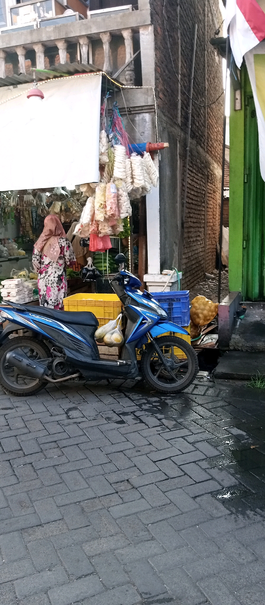The image depicts an outdoor scene in Wadungasri, Indonesia, possibly a market or street vendor stall. A blue and black Honda Vario scooter is parked on a wet, grey brick-paved street. The scooter has two bags of yellow fruit hanging from its handlebars. Behind the scooter, a woman wearing a pink headscarf and a floral-patterned dress is standing in front of a stall. The stall displays a variety of goods, including white and colorful packaged snacks hanging from the awning, and green vegetables wrapped in plastic. A yellow plastic crate is visible behind the scooter, and a blue plastic crate is to the right of it. In the background, a brick building with a balcony and some white awnings is visible. The environment appears to be slightly damp, suggesting recent rain.