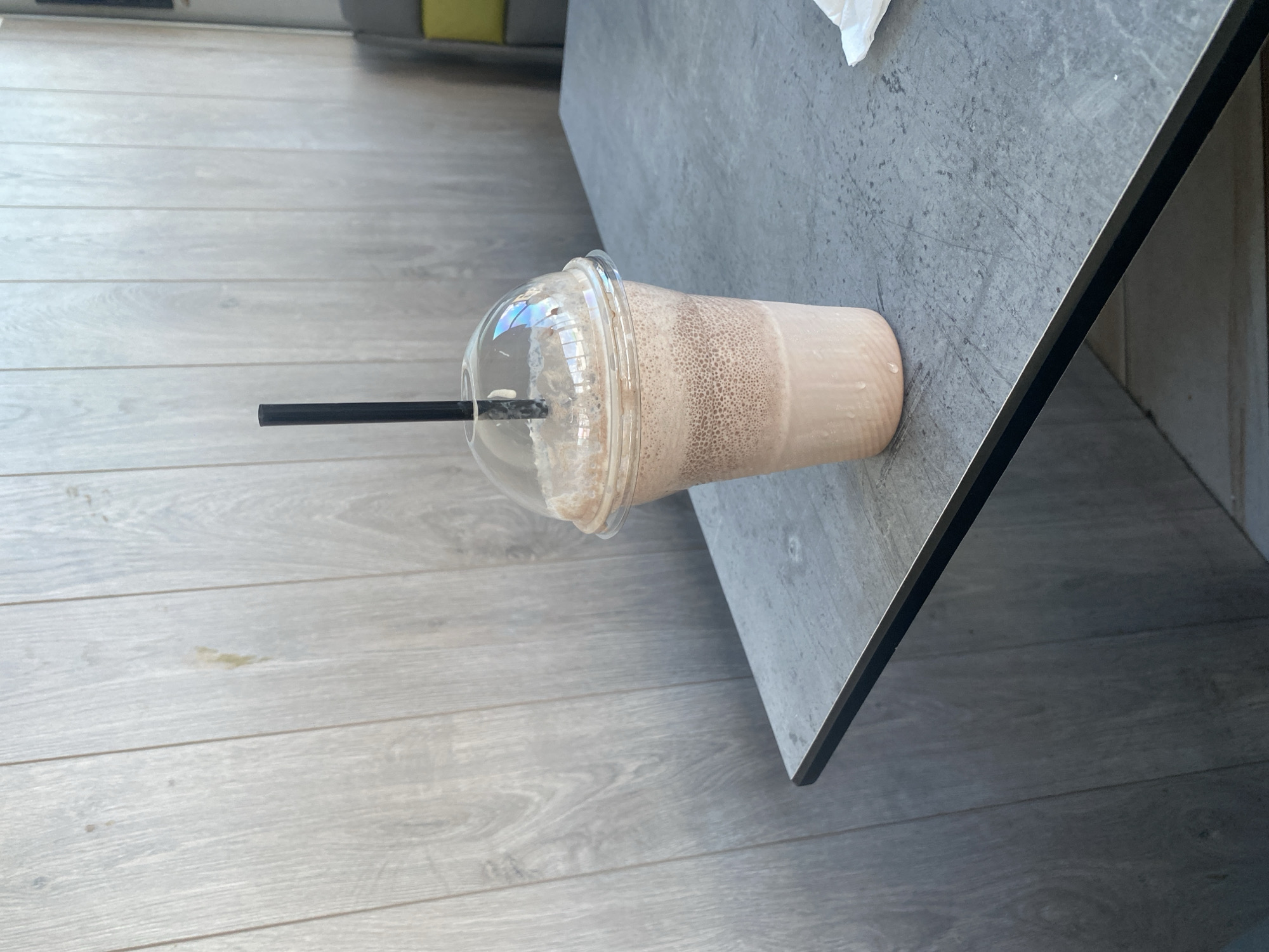 A close-up shot shows a chocolate milkshake in a clear plastic cup with a dome lid and a black straw, placed on a grey, textured table. The milkshake has a frothy top and appears to be a light brown color. The background is a light-colored wooden wall with vertical planks, suggesting an indoor setting. The table has a dark edge and is slightly angled in the frame. A white napkin or paper is visible on the right side of the table. The lighting suggests it is daytime. The location is identified as Ablanitsa, Bulgaria. There are no people or animals in the image.