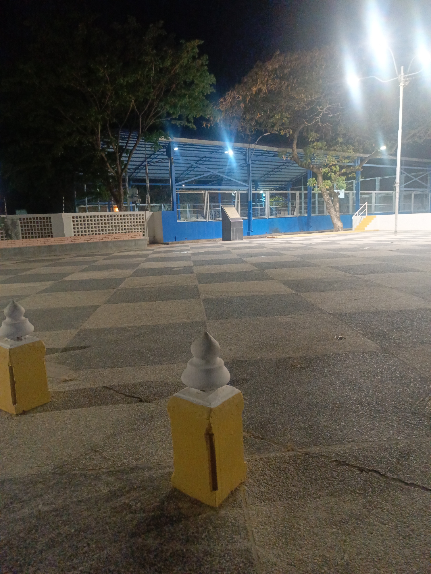 The image depicts an outdoor public space at night in Altagracia, Venezuela. In the foreground are two yellow, square pillars with white decorative tops. The ground is covered in gray and black checkered pavement.
Behind the pillars is an open-air basketball court enclosed by blue metal fencing. There are trees visible on either side of the court. A tall street light is situated on the right side, casting a bright glow over the area. There is also a short brick wall towards the left side of the court. The scene is illuminated by the streetlights and the court's lights, creating a somewhat dim environment. There is no indication of people present.