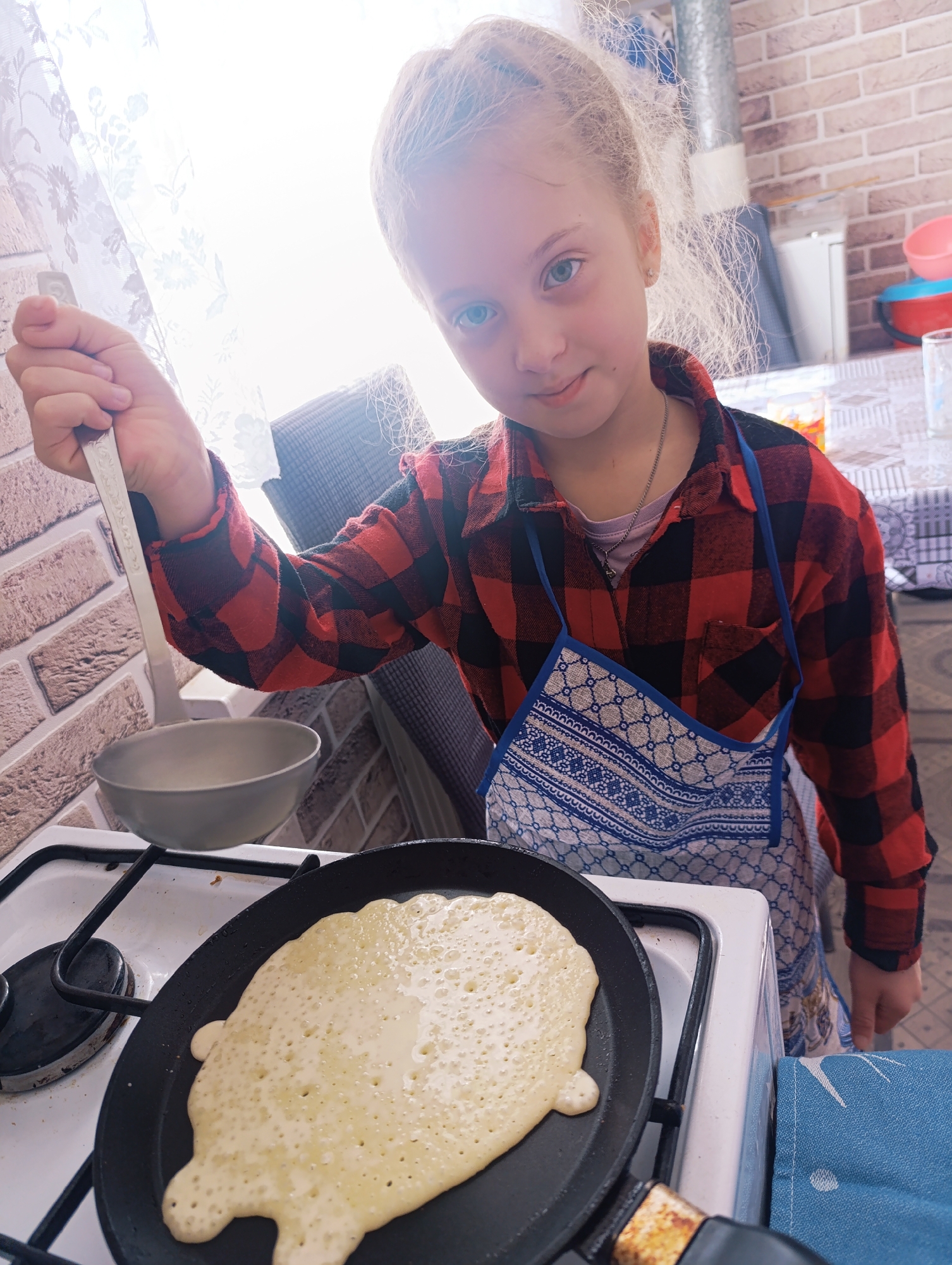 A girl with fair hair and blue eyes, wearing a red and black plaid shirt and a blue apron, is cooking pancakes in a kitchen. She is holding a metal spoon or ladle in her right hand. A pancake is in a black frying pan on a white stove top. To the left of the stove, there is a metal bowl. In the background, there is a window with a patterned curtain. The environment seems to be an indoor kitchen setting. The city and specific location are unknown.