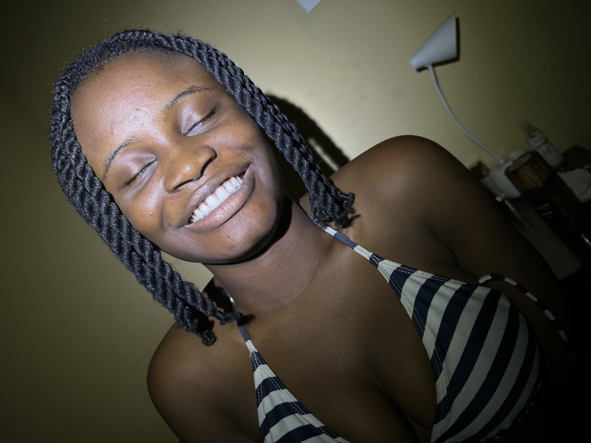 A high-angle, medium shot shows a Black woman with her eyes closed and a wide smile, revealing her teeth. She is lying down and looking upwards. She has dark, braided hair. She is wearing a blue and white striped bikini top. The background is a plain, light brown wall. A white desk lamp is visible in the lower right corner of the image. The scene appears to be indoors, possibly in a bedroom or living area. The lighting suggests it may be nighttime or a dimly lit room. The image does not provide specific details about the location in Ihiagwa, Nigeria, other than its general indoor setting.