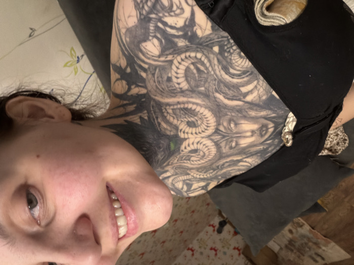A smiling person tilts their head towards the camera, revealing a large, detailed tattoo of Medusa on their upper arm and chest. The tattoo features intricate snake patterns and a striking depiction of the mythological figure. The individual wears a black top or bra. The background shows a patterned surface and a gray pillow. The lighting is indoor and somewhat diffused.