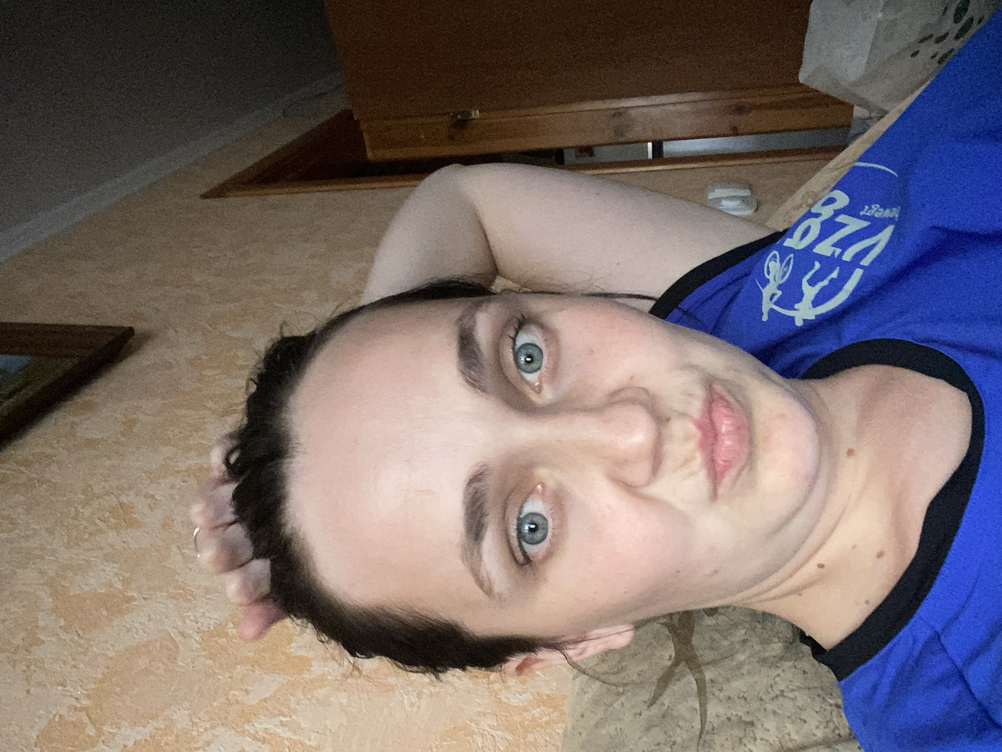 The image is a close-up selfie of a woman with dark hair pulled back, wearing a blue t-shirt. She has light blue eyes and is making a duck face. Her right arm is raised and bent behind her head. The background shows a textured wall and a wooden doorframe. The image is taken indoors. The scene is not identifiable as being in any specific city or location.