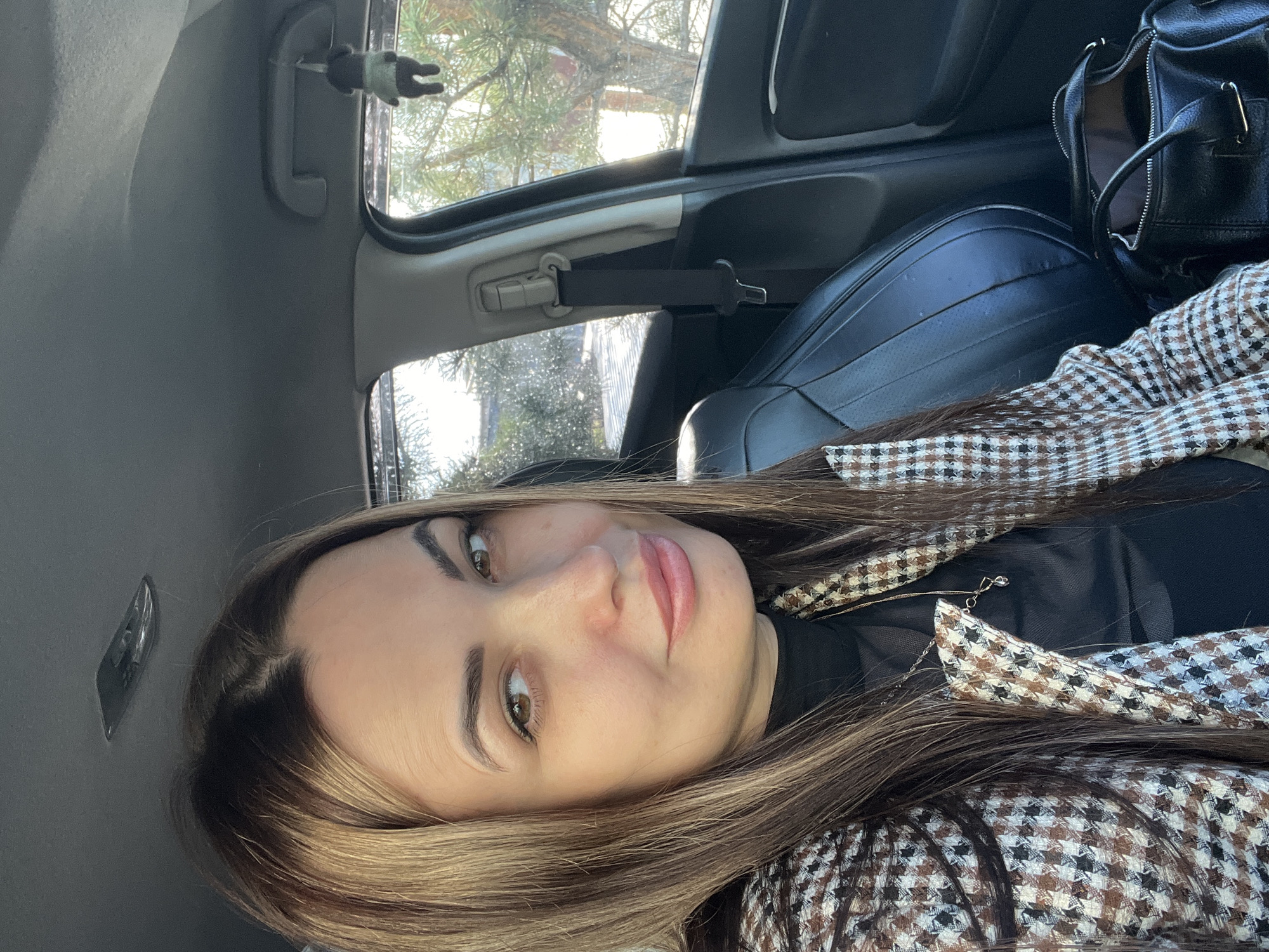 A selfie of a woman with long brown hair, wearing a black turtleneck and brown and white checkered coat, taken inside a car. A black purse is on the seat beside her. The car is parked with a view of trees and possibly a building outside the window.