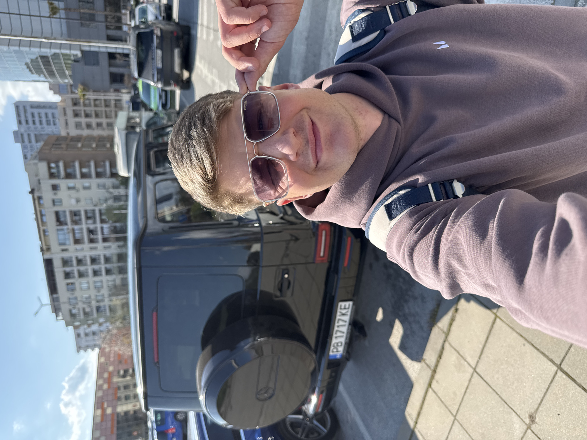 A man takes a selfie outdoors on a sunny day in an urban setting, likely Plovdiv, Bulgaria, given the visible license plate. The man, with light-colored hair, is positioned on the right side of the frame, looking directly at the camera with a relaxed smile. He wears silver-framed rectangular sunglasses and a brown or mauve hooded sweatshirt, featuring a small white, stylized logo (resembling two curved lines) on the chest. His right hand is visible near his head, possibly adjusting his sunglasses or hair.
Behind the man, a black Mercedes-Benz G-Wagen SUV is prominently parked. Its spare tire cover is visible on the rear door, and its Bulgarian license plate, "PB 1717 KE," confirms the location as Plovdiv. The ground beneath the car and the man is a mix of light-colored sidewalk tiles and darker asphalt, indicating a street or parking area.
In the background, under a clear blue sky, an urban landscape unfolds with various buildings, including modern structures with glass reflections and older, multi-story residential or commercial buildings. The bright sunlight casts distinct shadows, suggesting it is likely mid-morning or afternoon. Other vehicles are partially visible in the distant background. The overall scene depicts a casual moment in a bustling city environment.