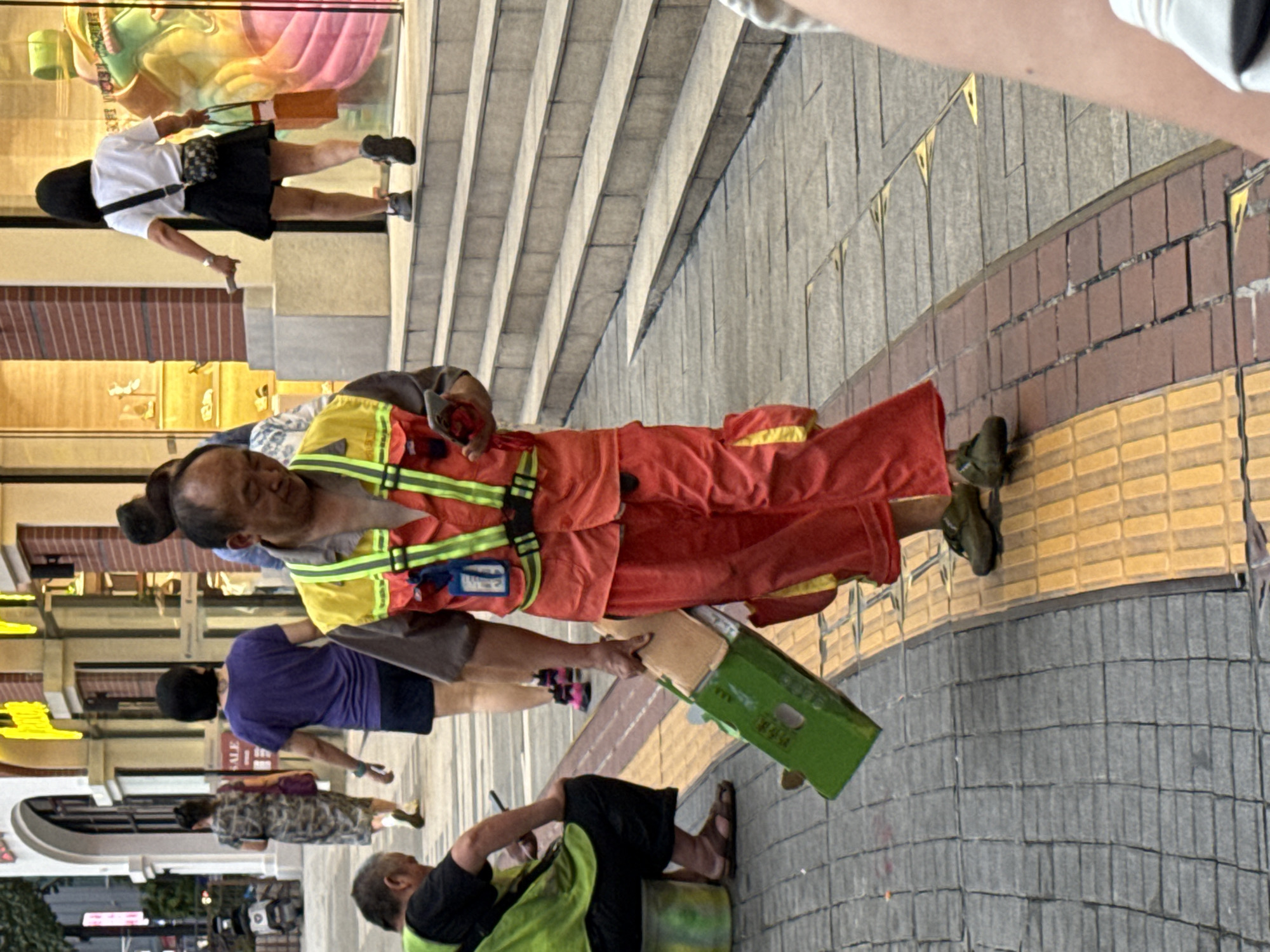 An older East Asian man, wearing a bright orange uniform with a yellow and green reflective vest, walks on a sidewalk in Yuzhong District, China, holding a green cardboard box at an angle. The man is looking down at his mobile phone, which he holds in his right hand. His uniform includes bright orange pants with a yellow pocket. In the background, other pedestrians are visible, some walking on stairs and others on the sidewalk. One woman in the background is wearing a black skirt and white t-shirt and carrying shopping bags. The sidewalk is paved with grey and yellow textured tiles. Buildings with storefronts are visible behind the pedestrians. A motorcycle is parked on the left side of the image.