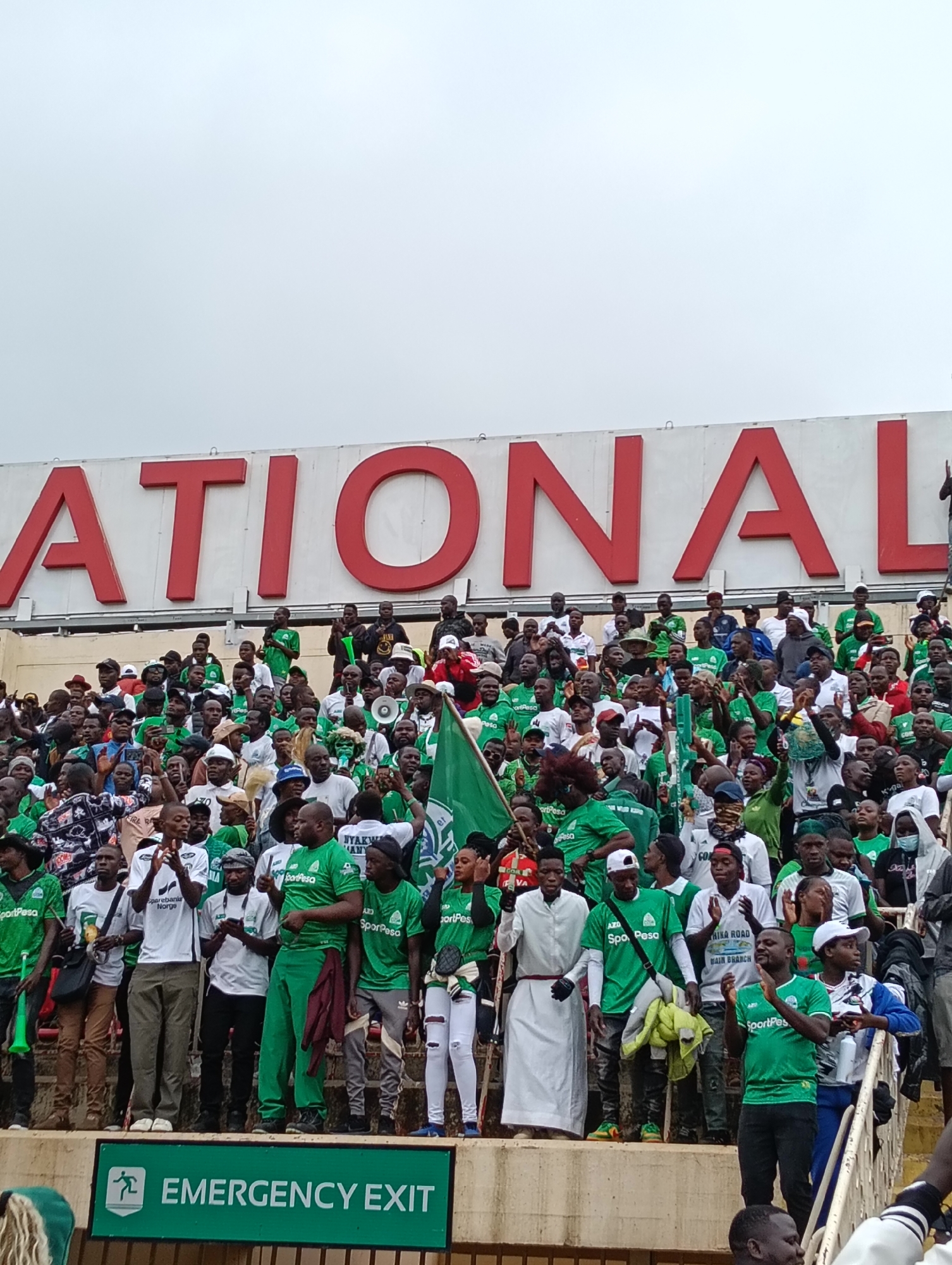 The image shows a large crowd of people at a stadium, with the word "NATIONAL" prominently displayed in large red letters on a white background above them. The crowd appears to be mostly composed of men and women, many of whom are wearing green and white clothing, suggesting they are supporters of a sports team. Some individuals are holding flags and engaging in actions typical of a sporting event, such as clapping and cheering. The environment appears to be an outdoor stadium setting with tiered seating. Below the crowd and to the right, an "EMERGENCY EXIT" sign is visible in green and white. While the image is taken in Nairobi, Kenya, the specific stadium is not identified.