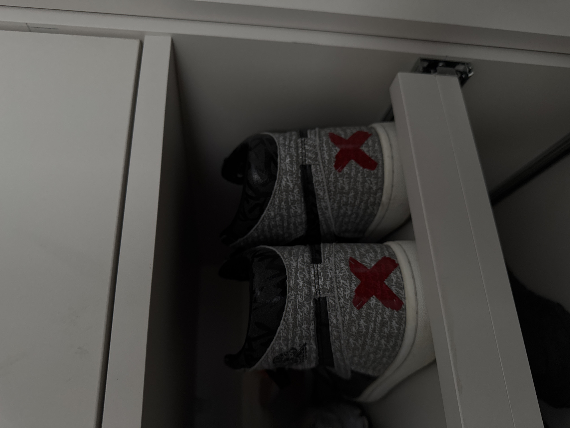The image displays a static, indoor scene within a light-colored storage unit, identified as being in Minsk, Belarus. Two high-top sneakers are centrally positioned, resting side-by-side on a white pull-out shelf or drawer. The sneakers feature a textured, light gray upper material with subtle, dark script patterning, black ankle collars, and white outsoles. A distinct, hand-painted or applied red 'X' mark is visible on the heel counter of each shoe. Above the footwear, another light-colored horizontal surface, possibly a shelf or the bottom of an upper compartment, is visible. To the right, a portion of a metal drawer slide mechanism is discernible, attached to a vertical white panel. No individuals or active interactions are present in the scene.