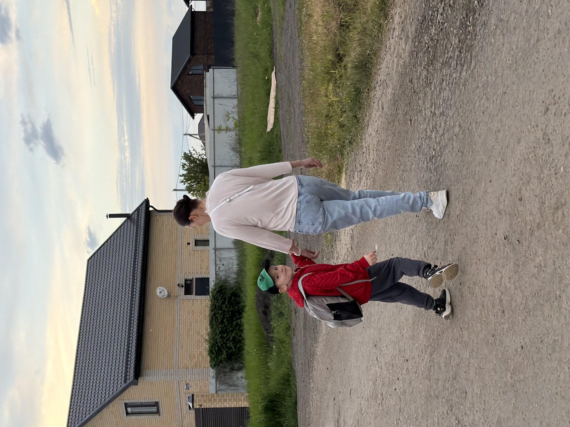 The image depicts a woman and a boy walking on a dirt road in what appears to be a residential area. The boy is wearing a red jacket, gray pants, black and gold sneakers, a green baseball cap, and a gray backpack. The woman is wearing a long-sleeved white shirt, blue jeans, and white sneakers. The boy is holding a white object in his right hand. The location is in Rossiyskiy, Russia. There are houses and green vegetation along the roadside. The sky is visible with some clouds.