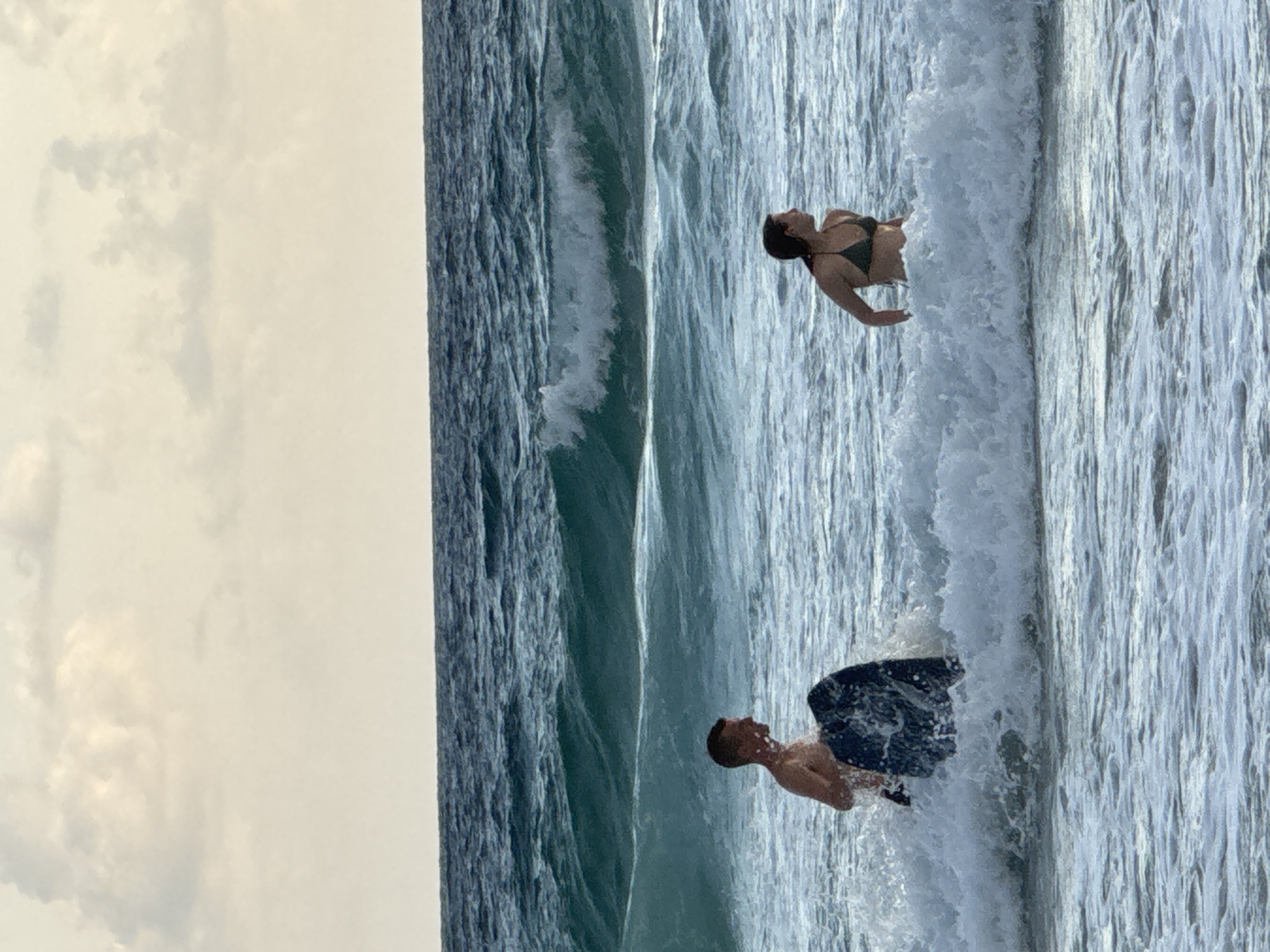 The image shows two people in the ocean off the coast of Karon, Thailand, with waves breaking around them. On the left, a man is partially submerged in the water, holding a dark-colored boogie board. He is looking to his right. On the right, a woman is also in the water, wearing a dark green bikini. She is looking to her right. The sea is a deep blue-green, with white foam from the waves. The sky above is overcast with light-colored clouds.