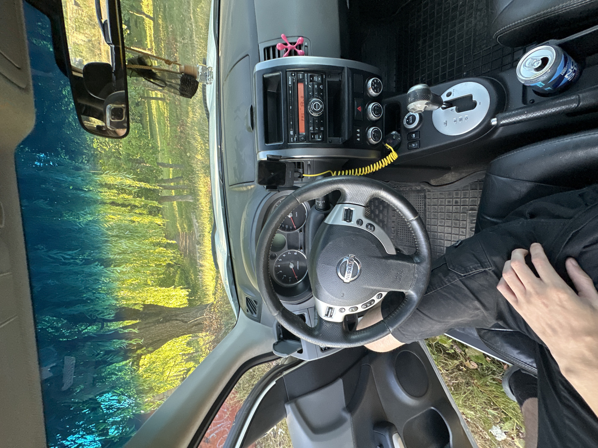 The image shows the interior of a car, likely a Nissan X-Trail, seen from the driver's perspective. The car is parked outdoors in a grassy area with trees. The windshield reflects the light blue sky and the foliage of willow trees. A person's legs, clad in black pants, are visible in the driver's seat, with their right hand resting on their thigh. The dashboard features a steering wheel with a Nissan logo, a radio, and climate controls. A mobile phone is mounted on the dashboard. The gear shift is in the "P" (Park) position. A can of energy drink is placed in the center console. The scene appears to be in Makiv, Ukraine, given the context provided.