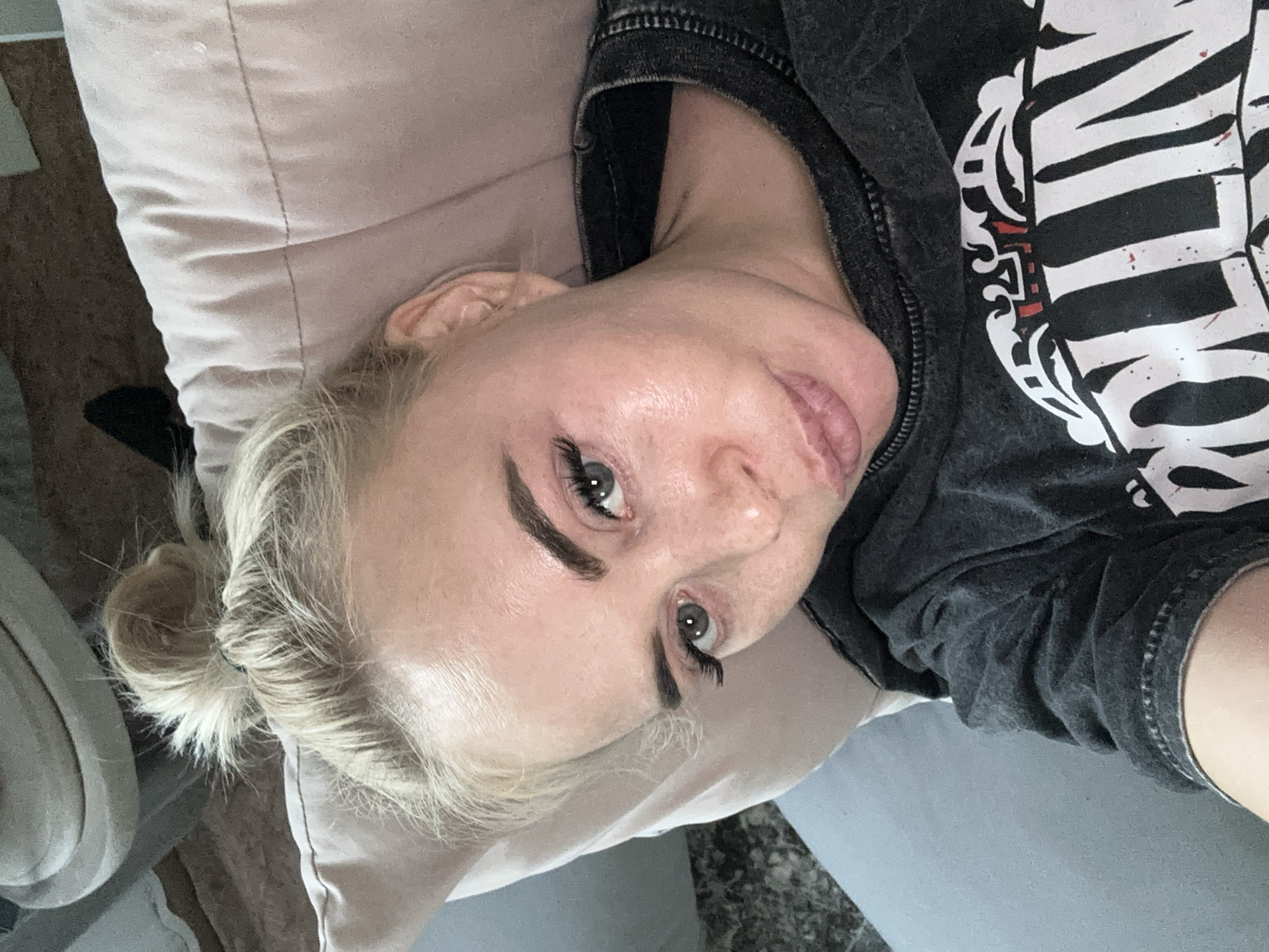 The image shows a fair-skinned person with blonde hair pulled back in a bun, lying on a light pink pillow and a gray sofa. The person is wearing a dark gray t-shirt with a white and black graphic print. The person's face is turned towards the camera. The location is unknown and there is no evidence of the city of Высокий, Russia.