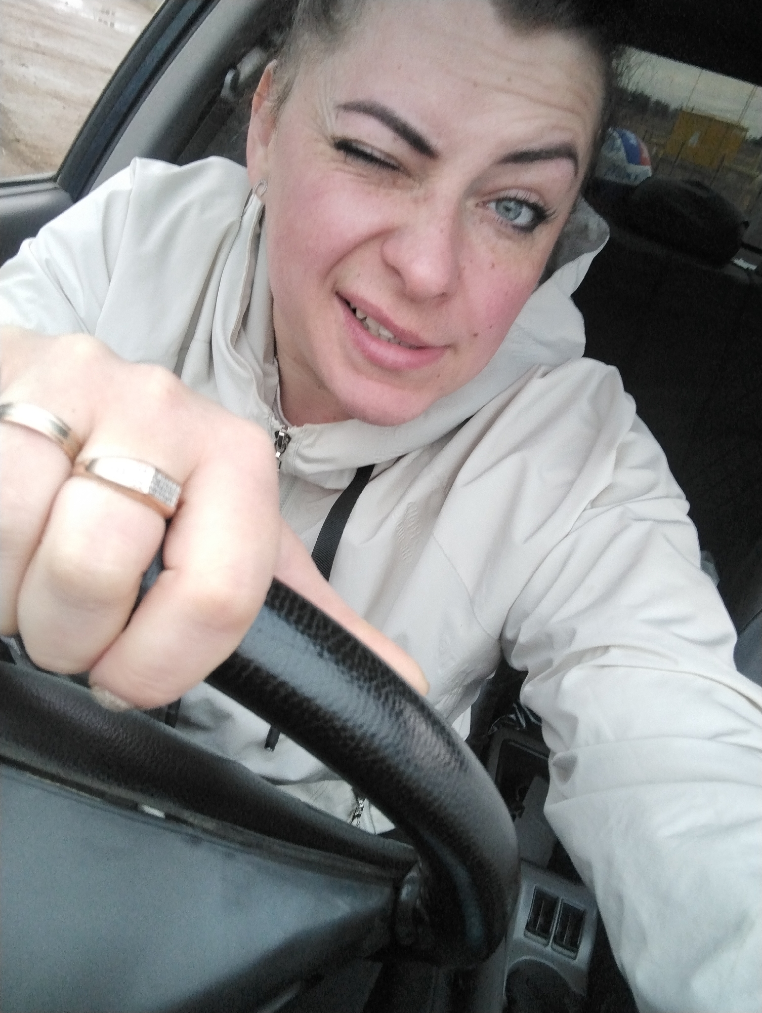 A light-skinned woman with blue eyes is winking while sitting in a vehicle, likely the driver's seat, as indicated by her hand on the steering wheel. She is wearing a light-colored jacket and rings on her fingers. The background shows elements of an outdoor environment and the interior of the vehicle. This image may have been taken in Grishanki, Russia.