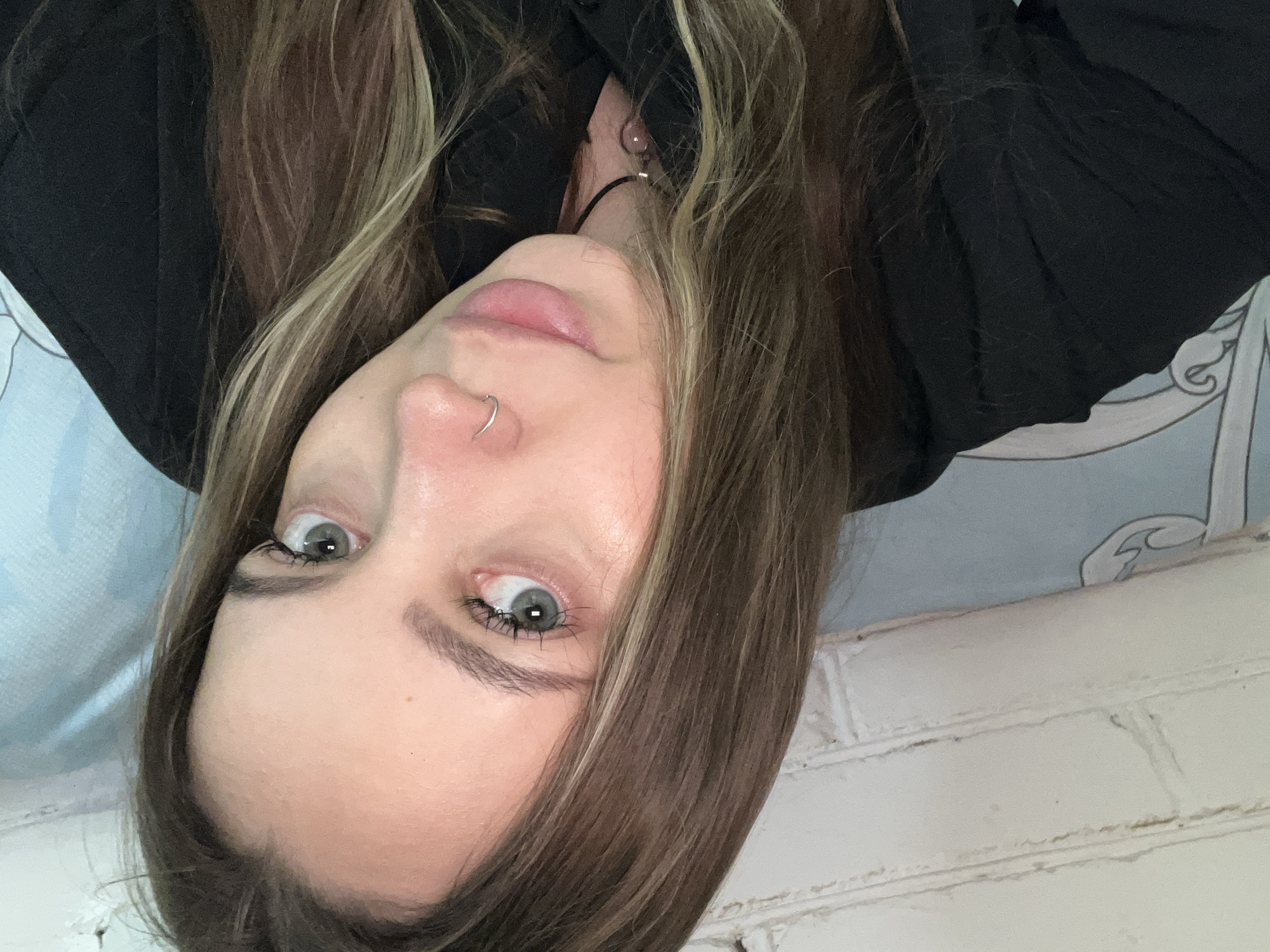 A selfie depicts a woman with light skin, light eyes, and light brown hair with blonde highlights. She has a silver nose ring. She is wearing a black shirt. The background appears to include a white brick wall on the bottom and a blue patterned surface on top. There is no clear evidence in the image to suggest this photo was taken in Novopokrovskaya, Russia.