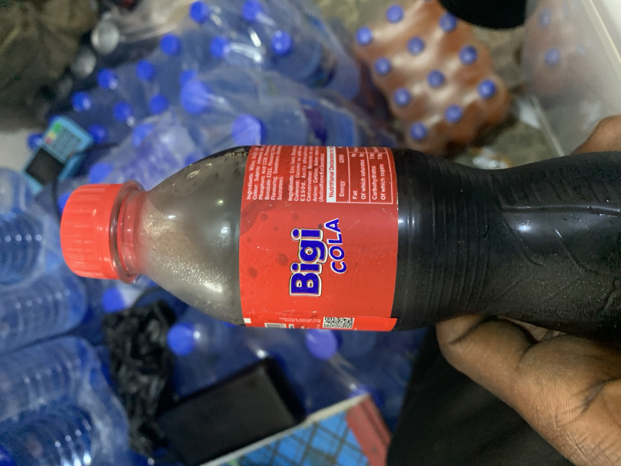 The image shows a close-up of a bottle of "Bigi Cola" held in a person's hand. The bottle is dark brown with a red cap and a red label that prominently displays the brand name. The label also contains nutritional information and ingredients in English and French, including details about water, sugar, colorants, acids, and sweeteners. Behind the bottle, there are numerous other bottles, many of them appearing to be water bottles with blue caps, stacked in the background. Some of these bottles are partially visible and appear to be arranged in a manner suggesting they are part of a larger display or stock. A black plastic bag is also visible on the left side of the frame, near the bottles. The overall environment suggests a retail or storage setting, possibly a shop or a warehouse. The presence of the "Bigi Cola" brand, which is a popular Nigerian beverage, strongly indicates the location is within Nigeria. Specifically, the bottle itself with its labeling and the surrounding context of what appears to be a large quantity of beverages are consistent with a setting in Nigeria. New Stadium, Nigeria, is a locality, and this scene could plausibly be found in a commercial establishment within such an area, such as a local supermarket, a beverage distribution center, or even a roadside vendor.