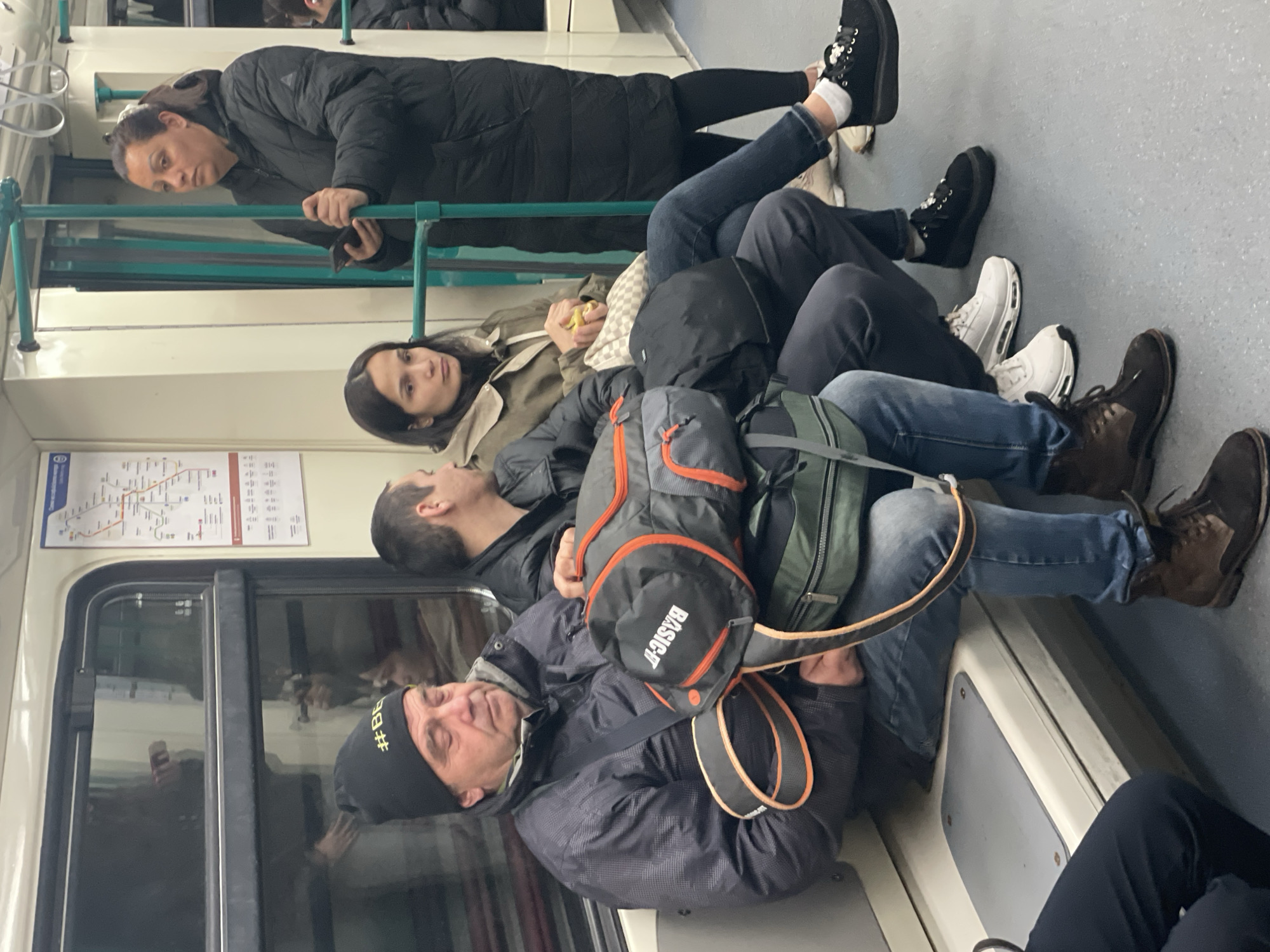 The image captures a group of people riding on a train. The interior of the train is visible, with windows, seats, and a route map. The individuals are diverse, with different ages, genders, and clothing styles. One woman is standing and holding onto a rail, looking at her phone. Other people are seated, and some appear to be resting or sleeping. The environment is that of a public transportation setting. Based on the appearance of the train, the map on the wall and some visible graffiti, the location may be the Sofia Metro subway system in Sofia, Bulgaria.