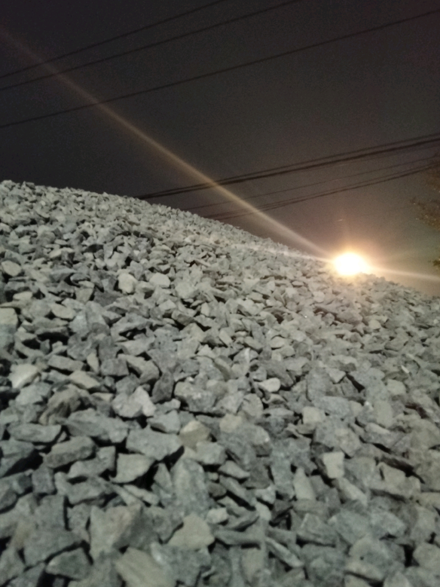 The image depicts a large pile of gray gravel under a dark sky at night. Several dark, horizontal power lines are visible in the upper portion of the frame. A bright light source, possibly a streetlamp or vehicle headlights, is causing a lens flare and illuminating the gravel from the right side, casting a warm glow and a light streak across the scene. The perspective is low, looking up at the pile of gravel, which fills the foreground and extends into the mid-ground. The gravel appears to be a construction material. There are no people or animals visible in the image. The location in Pontianak, Indonesia is not discernible from the visual information provided in the image itself.