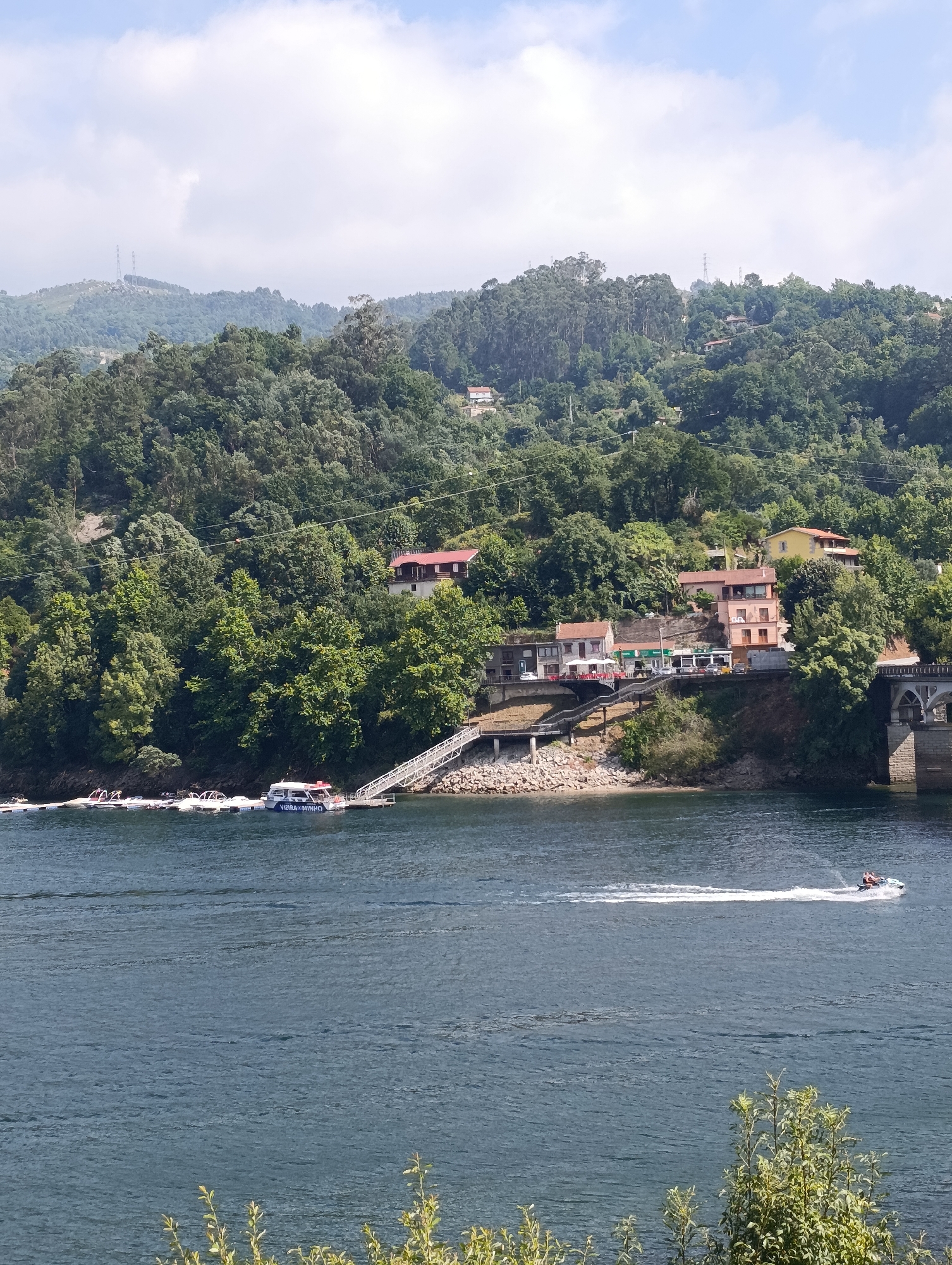 This image depicts a scenic view of the Cávado River in Terras de Bouro, Portugal. The river, with its dark blue, rippling water, dominates the foreground. In the middle ground, a tree-covered hillside rises steeply from the riverbank. Several buildings, including houses and shops, are nestled into the slope of the hill, some with red-tiled roofs. A bridge with stone arches spans the river on the right side of the image.
On the river, a white passenger boat with "VIEIRA MINHO" written on its side is docked at a pier with a ramp leading to the shore. Further out on the water, a jet ski with two people on board is creating a white wake as it speeds across the surface. The background features more hills covered in dense green trees, with a few power lines visible traversing the landscape. The sky above is a mix of blue and white clouds.