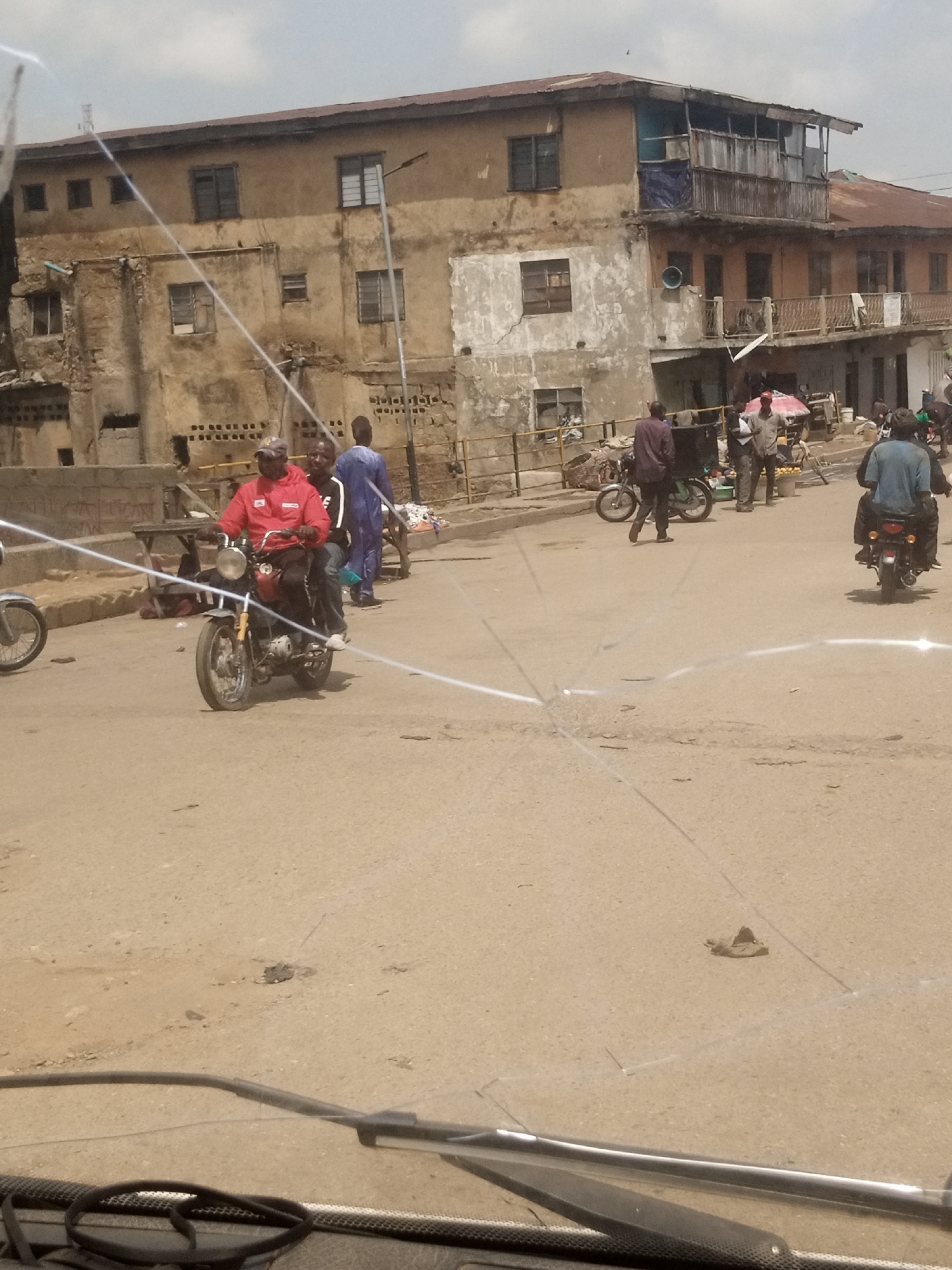 The image depicts a street scene in Jos, Nigeria, under a cloudy sky. In the foreground, the cracked windshield of a vehicle offers a blurred view of the road ahead. A person in a red jacket is riding a motorcycle, accompanied by a passenger. To the left of the motorcycle, a second motorcycle is visible, and to the right, another motorcycle is seen from the rear with its rider.
In the background, a weathered, multi-story building with an upstairs balcony and a rusty roof dominates the scene. Several individuals are standing on the street, and a cart with baskets is present. A street lamp is mounted on a pole near the building. The ground is paved with asphalt and shows signs of wear and tear, with some debris scattered about. The overall impression is one of a bustling, ordinary day in a developing urban environment.
