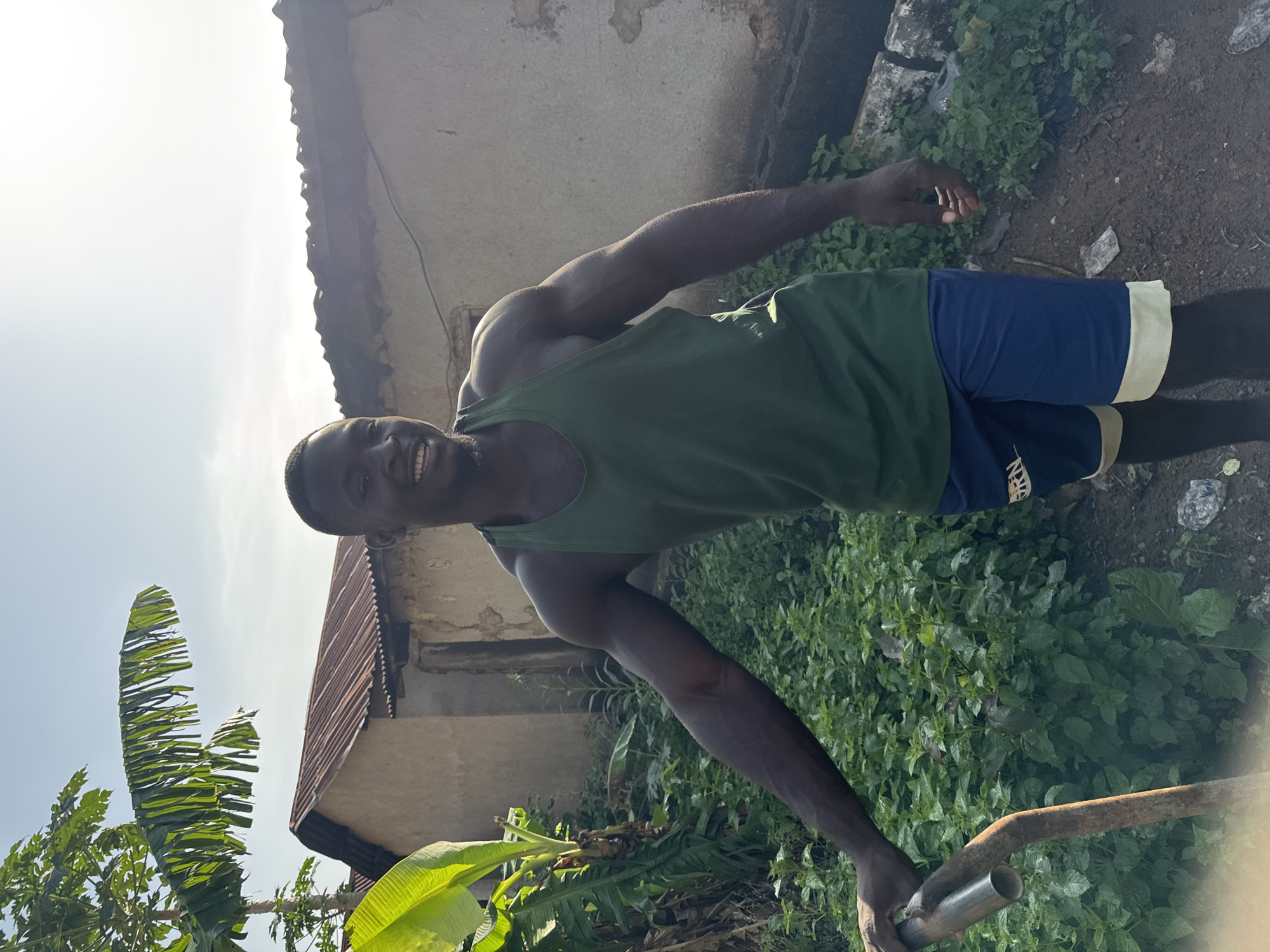A dark-skinned man wearing a green tank top and blue shorts with yellow trim stands in a yard in Akwanga, Nigeria, holding a rusty metal tool. He has a wide smile and muscular arms. Behind him, a building with a corrugated metal roof and a concrete wall is visible, along with lush green vegetation, including banana trees. The sky is bright and clear.