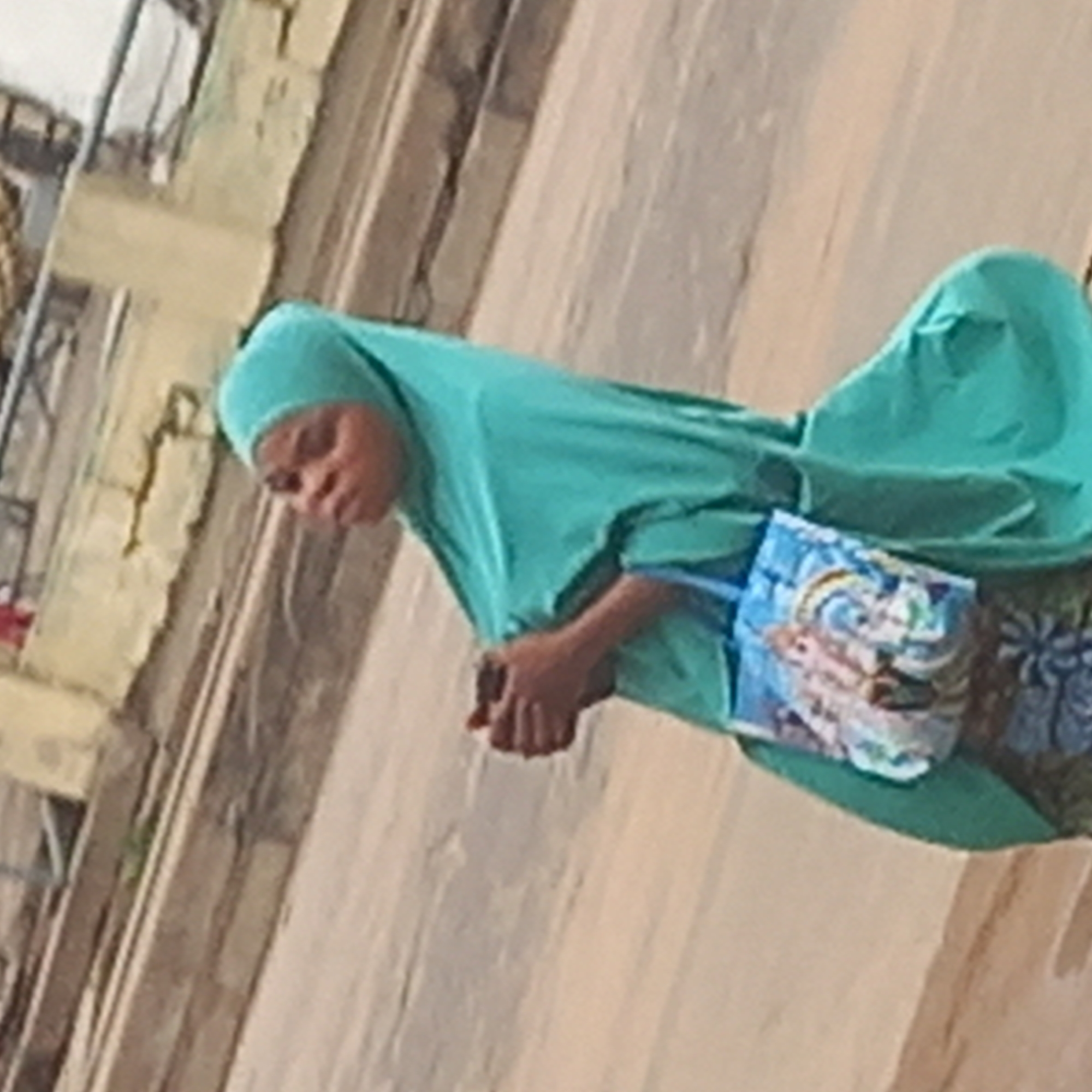 This tilted, somewhat blurry daytime image captures a woman standing or walking outdoors, possibly in Dadi Riba, Nigeria. The woman, who appears to be of African descent, is dressed in a teal-colored head covering (hijab) and a matching, loose-fitting outer garment that drapes over her body. Her hands are clasped in front of her, and she carries a small light blue shoulder bag adorned with a colorful, illustrative design featuring what appears to be a unicorn or fantasy creatures among clouds. A patterned fabric, possibly a skirt or dress, is visible beneath her outer garment. Her facial expression is neutral or contemplative. The background consists of a worn, light-colored building wall or structure, showing some texture and what may be dirt streaks, and a rustic, unpaved ground surface. There is no discernible text or specific activity beyond her presence in the outdoor setting.