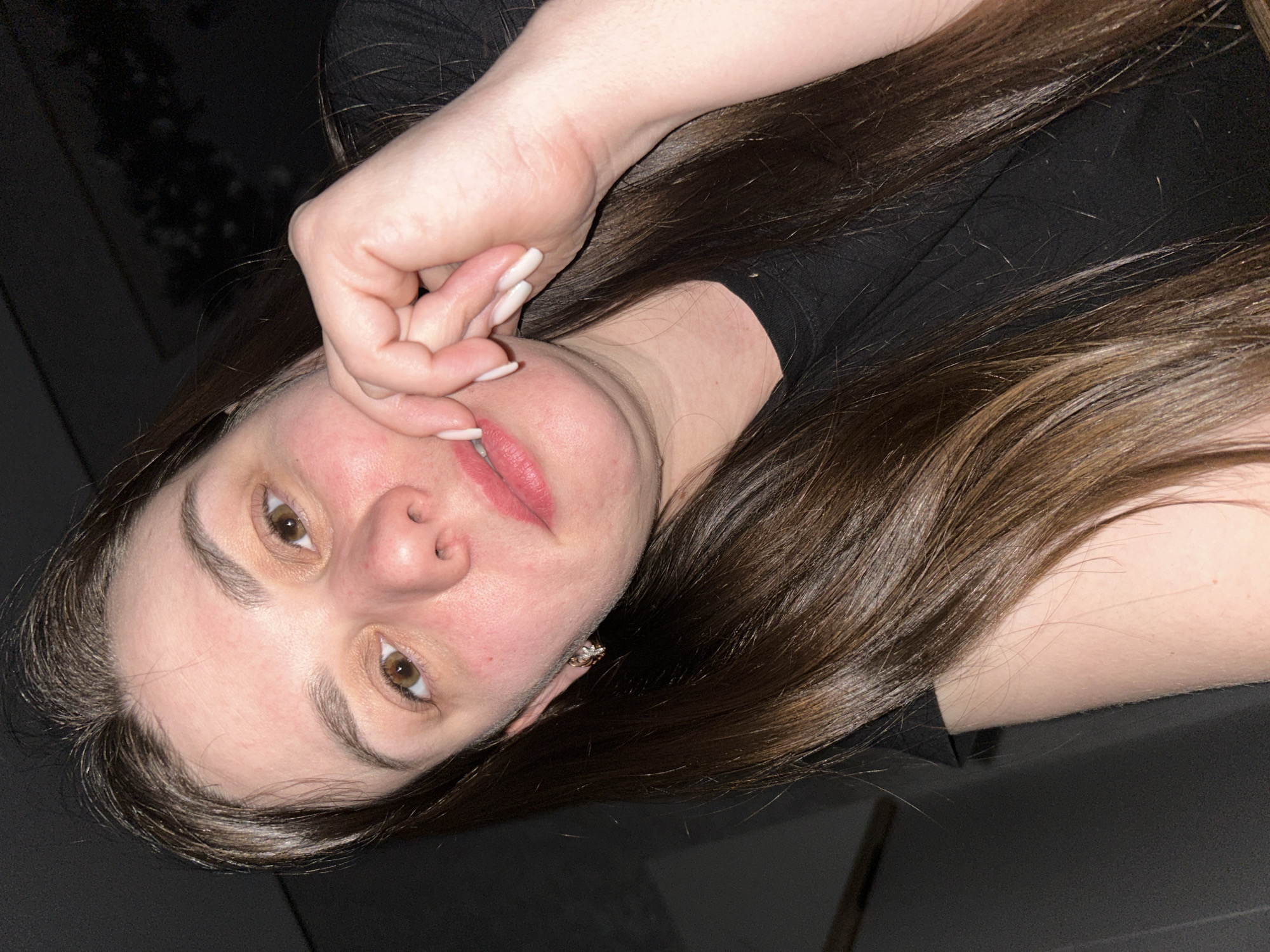 A medium close-up shot depicts a woman with light skin and light brown hair. She is wearing a black shirt. Her face is slightly flushed, and she has brown eyes and pink lipstick. Her right hand is raised, with her fingers near her face. The location and city of Chaikovsky, Russia cannot be determined from the image alone.