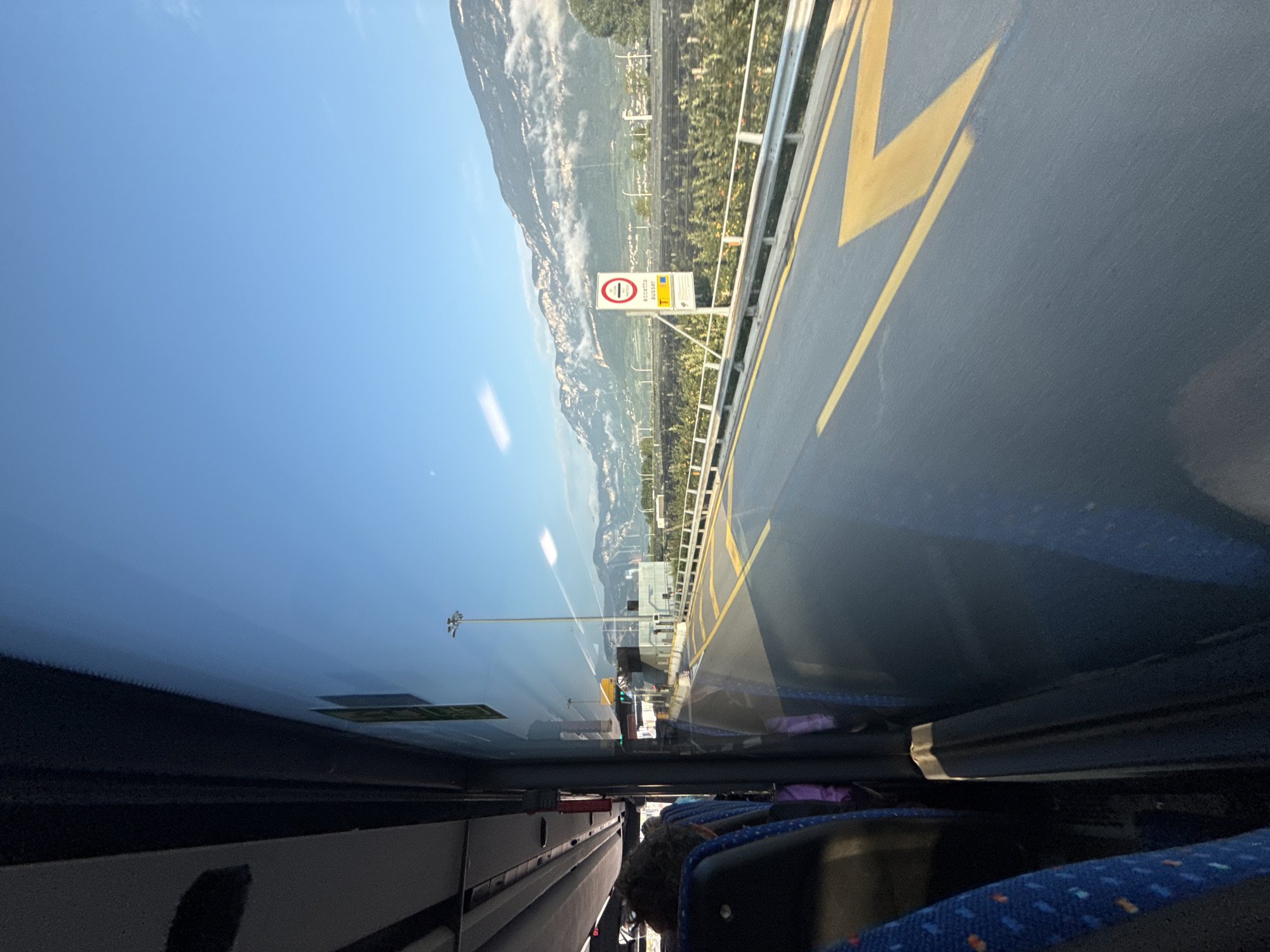This is a photo taken from inside a moving bus on a clear, sunny day. The bus is traveling on a road with yellow lane markings. In the distance, there are mountains partially covered by clouds. A road sign is visible on the right side of the image, depicting a circular red sign with a horizontal black bar inside, indicating a prohibition, and below it, text that appears to read "eccetto estero" and "T". This suggests the location might be near a border crossing, potentially indicating a toll or specific entry requirements for foreign vehicles. The scenery suggests a location in or near an Alpine region.