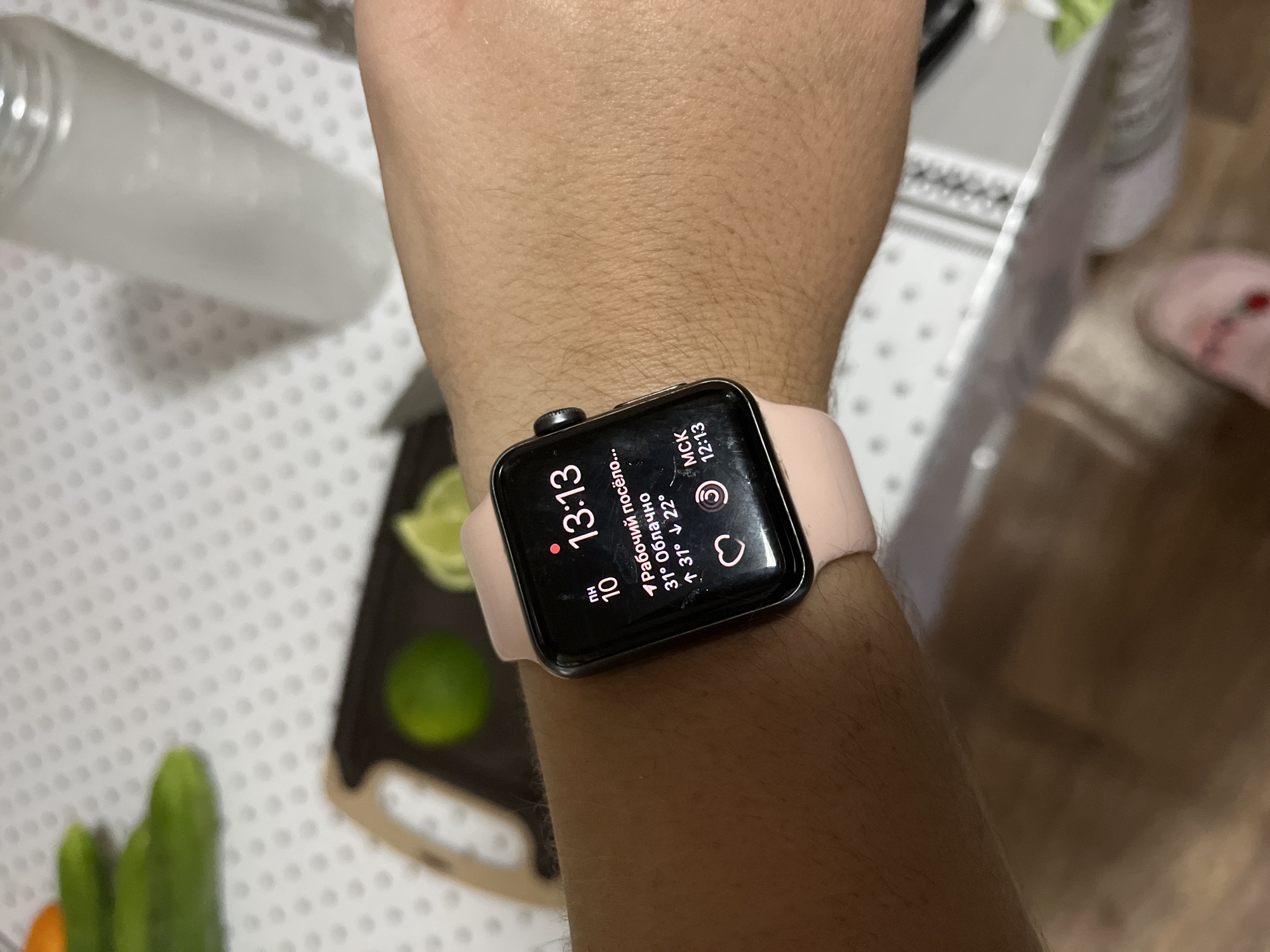 A close-up shot captures a person's wrist wearing a pink Apple Watch. The watch face displays the time as 13:13, the date as the 10th, and a weather forecast indicating "partly cloudy" with temperatures of 31 degrees Celsius rising and 22 degrees Celsius falling. Below the weather information, there's a heart icon and a circular icon, possibly representing activity tracking. The watch is positioned on a person's arm, which has visible hair. In the background, out of focus, are kitchen items including cucumbers, limes, a cutting board, and a partially visible water bottle. The scene appears to be indoors, likely in a kitchen environment within Krasnyye Barrikady, Russia.