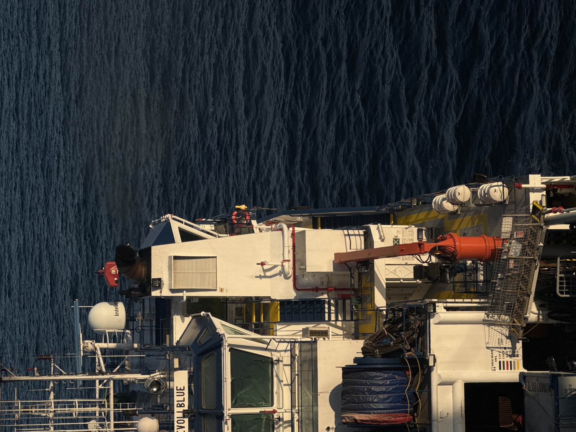 An eye-level, long shot captures a portion of a white research vessel named "EVOLBLUE" against a dark, choppy sea. The ship's superstructure, predominantly white with blue accents, dominates the left and central portions of the frame. Various deck equipment is visible, including a radar dome with the "Intelli" logo, a red horn-like structure, a white ventilation unit, and a small red crane. Yellow safety lines and staircases are also apparent on the deck. Multiple life rafts are stacked on the right side of the ship. The sea is a deep, uniform blue, reflecting the clear, daytime sky, and appears to be in motion with small waves. The lighting suggests it is daytime, possibly morning or late afternoon, given the warm sunlight casting shadows on the vessel. No people are visible in the image. The location is unknown, and the description does not include any information about Italy.