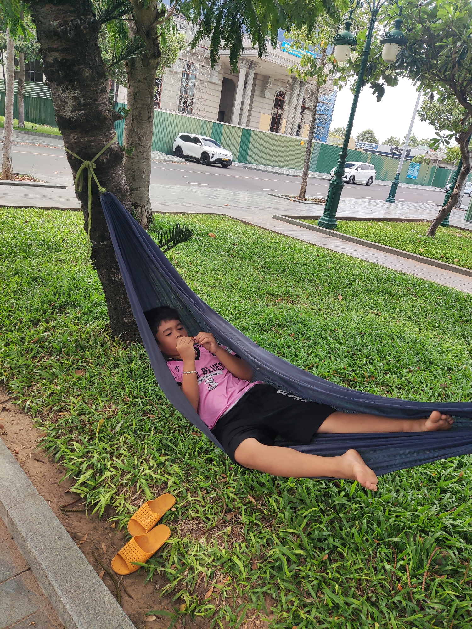 A young person lies in a hammock tied to a tree in an urban park setting. The individual is dressed in a pink shirt and black shorts. A pair of yellow sandals rest on the ground near the hammock. The park features green grass and other trees in the background, with a sidewalk and street visible in the background. Cars are seen parked on the side of the street and a building is under construction in the background behind a green wall. The city is Unknown.