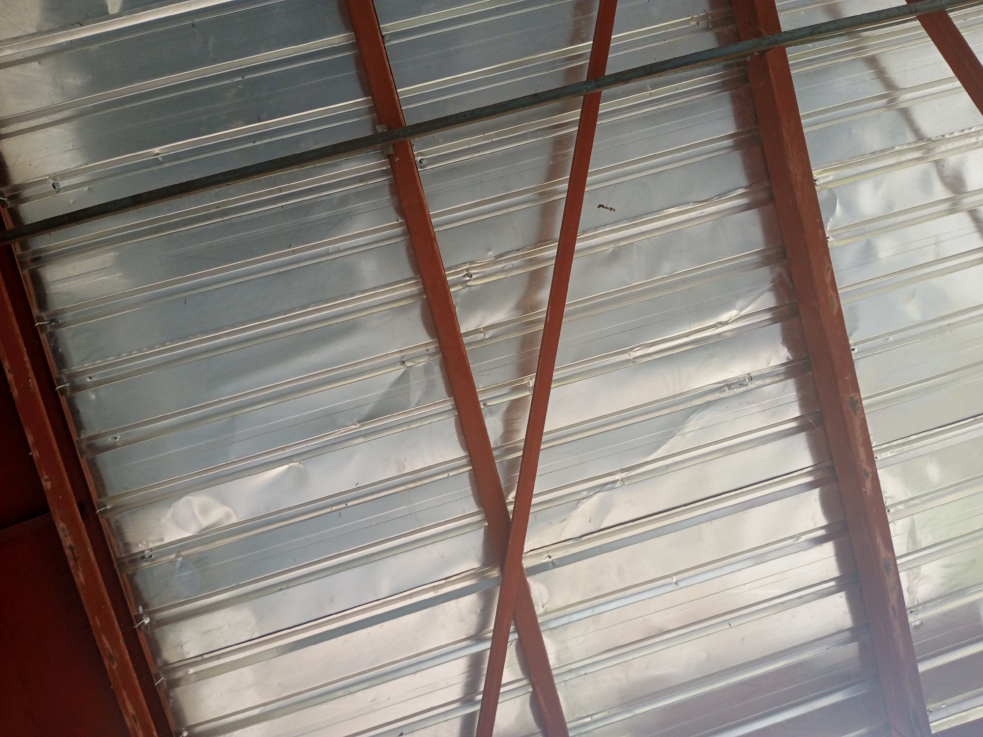 The image shows the underside of a corrugated metal roof, supported by reddish-brown metal beams. The roof panels are silver and have a ridged texture. Two of the metal beams are angled and cross each other near the center of the image, creating an "X" shape. A thin, dark horizontal bar runs across the top of the image, also made of metal. The overall impression is of an industrial or utilitarian structure, possibly a workshop or storage area. The lighting suggests it is daytime, with bright light reflecting off the metal. There are no people visible, and no text is present. The scene appears to be in Jalingo, Nigeria.