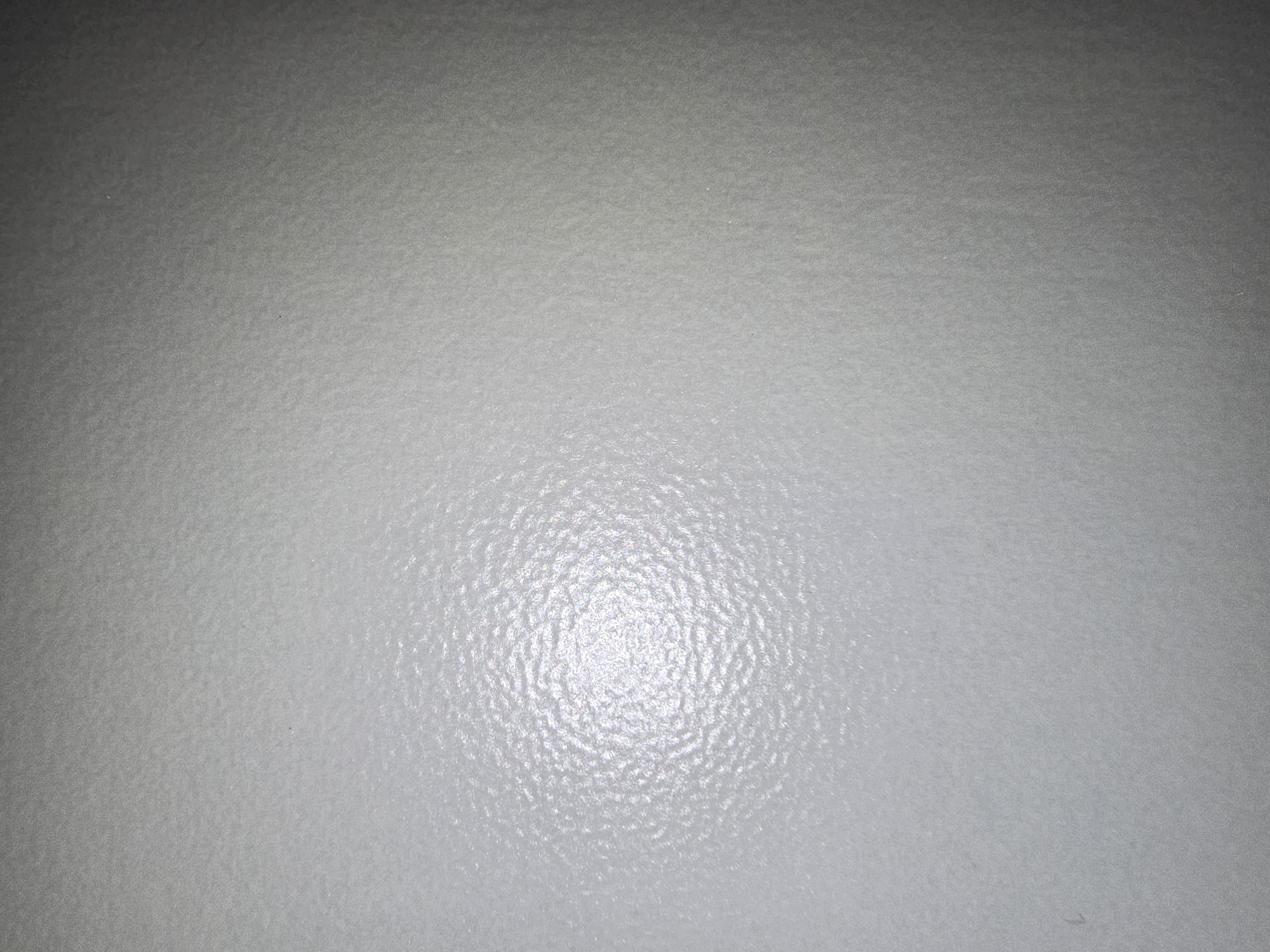 The image shows a close-up, abstract view of a textured, light gray surface. The texture is irregular, with small bumps and ridges that create a mottled appearance. There is a bright, diffused light source to the left of the frame, causing a strong highlight and a gradient of light and shadow across the surface. The overall impression is that of a common, man-made material, possibly a wall or a textured panel. There are no people, specific objects, or discernible environmental features that would allow for identification of a location within the city of Ulan-Ude, Russia.