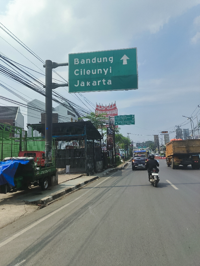 The image shows a street scene in Padalarang, Indonesia, at the intersection of Jalan Raya Bandung and Jalan Cileunyi. A large, green road sign with a white arrow pointing upwards reads "Bandung Cileunyi Jakarta." Below the sign, a smaller red sign with white lettering reads "Sayang Ibuah." Several other smaller signs are also visible. In the background, a black truck with a yellow cargo bed drives down the road. A light-colored car is visible in the distance. A black moped, driven by a person wearing a black jacket, is driving down the road. A green truck with a blue tarp covering the cargo bed is parked on the side of the road. The pavement is gray and shows signs of wear. There are several power lines strung above the road. Buildings and trees are visible on the sides of the road. The sky is blue and clear. The image was taken during the daytime, under bright sunlight.