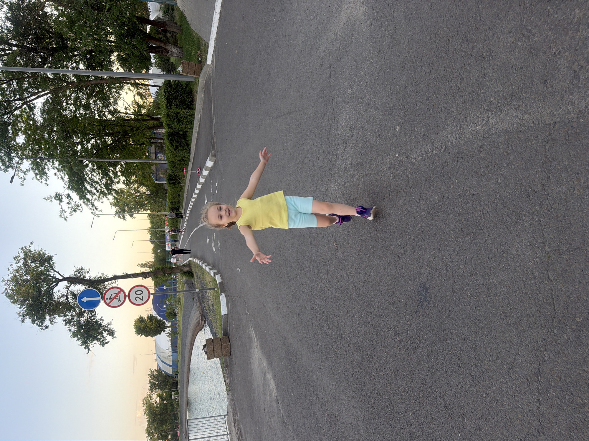 A young girl is lying prone on a gray asphalt road, her arms outstretched. She is wearing a yellow shirt, light blue shorts, and purple shoes. The road is adjacent to a body of water, potentially a lake or river, with a gray metal railing along the edge. Trees and greenery are visible on both sides of the road. Traffic signs indicating "No Motor Scooters" and a speed limit of "20" are nearby. This location is in Rostov-on-Don, Russia.