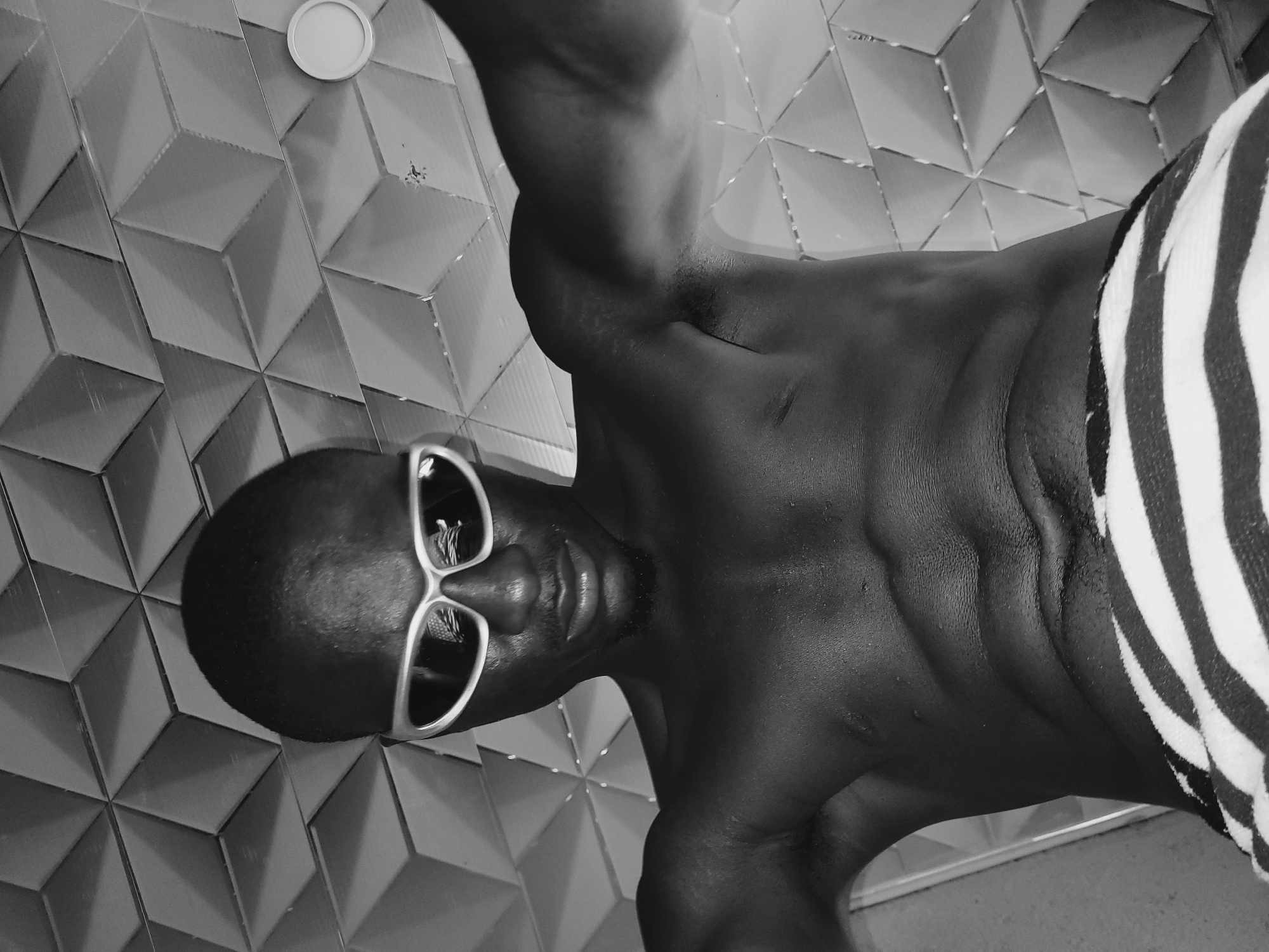 A black and white, vertically oriented medium shot captures a dark-skinned individual posing in what appears to be an indoor or semi-enclosed setting in Bukuru, Nigeria. The subject, seemingly male, is depicted from the waist up, with their torso and head occupying the foreground. They are wearing stylish white-framed sunglasses that reflect a distorted light source, obscuring their eyes. A slight stubble is visible on their chin.
The person's bare torso is prominently featured, showcasing defined musculature and glistening skin, suggesting recent exposure to water or sweat. A portion of a striped garment, possibly a towel or swim shorts, with bold black and white stripes, is wrapped around their lower body on the right side of the frame. The individual's head is tilted, and they are looking directly towards the camera, conveying a confident or relaxed demeanor.
The background consists of a textured wall covered in geometric tiles that create a repeating pattern of three-dimensional cubes or pyramids, adding depth and visual interest to the scene. A small, circular, light-colored fixture is visible on the wall in the upper left corner. The monochromatic palette emphasizes contrasts in skin tone, shadows, and the patterns of the tiles and garment. No text is visible in the image.