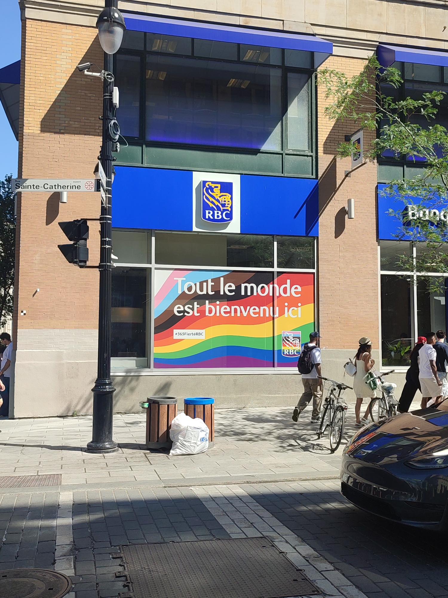 In Montreal, Canada, on a bright, sunny day, a scene unfolds on Sainte-Catherine Street. A prominent RBC bank branch features a large window displaying a pride flag-themed advertisement with text that translates to "Everyone is welcome here." The bank's blue signage is visible above the window, featuring the RBC logo.
In front of the bank, a black lamp post stands near a street sign indicating "Rue Sainte-Catherine Ouest." Two trash receptacles, one wooden and one blue, are placed near the sidewalk, with a large white bag of trash beside them.
Several people are walking along the sidewalk. A man with a backpack walks away from the camera, accompanied by a woman pushing a bicycle. Another woman, wearing a hat and a white dress, also pushes a bicycle. A group of people, including a man in a white t-shirt and shorts, walk towards the right side of the frame.
A dark-colored car, possibly a Tesla, is parked in the foreground, its front partially visible and reflecting the surrounding environment. The sidewalk is paved with grey bricks, and shadows from trees and structures are cast across the scene.