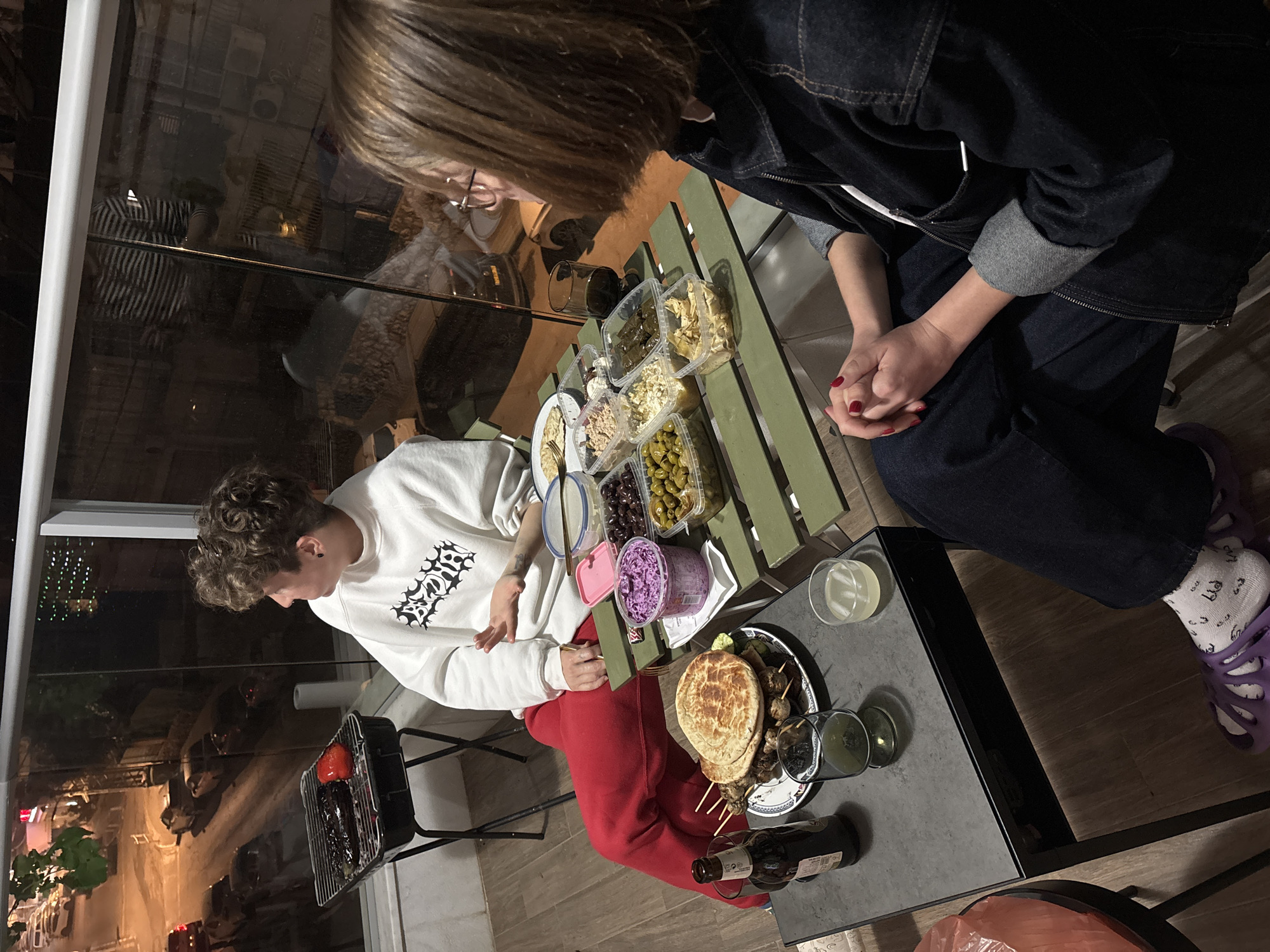 Two people are seated at two adjacent tables. One person wears a white sweatshirt and red sweatpants, and is seated to the left of the image. The other person, positioned to the upper right, wears a denim jacket and jeans, and has purple shoes.
The tables are covered with food items, including salads, pita bread, and skewers, along with drinks and a bottle of wine. A small grill is visible near the person in the sweatshirt, suggesting food preparation.
The image appears to be taken at night on a balcony, given the presence of a railing and the view of a city street, suggestive of Bat Yam, Israel. Reflections in the glass of the balcony can be seen.