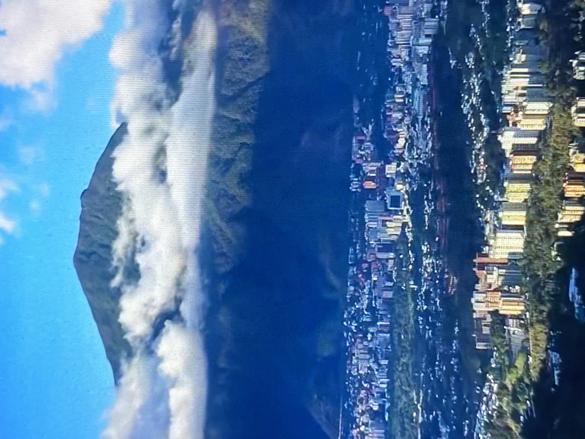 The image presents an aerial view, likely taken from an elevated position. On the left, a large mountain dominates the scene, partially obscured by clouds. The sky is blue, with scattered clouds near the mountain. Moving towards the right, the image reveals a city landscape, likely Tinaquillo, Venezuela. Buildings of various sizes and colors are visible, interspersed with trees and vegetation. The city appears densely populated, with structures closely packed together. The image is divided vertically with nature on the left and the city on the right.