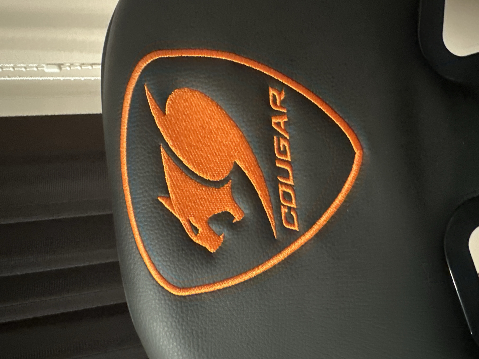 A close-up photograph captures a bright orange embroidered logo on a dark, textured surface, consistent with the backrest or headrest of an upholstered chair. The logo features a stylized cougar head facing right, with an open mouth, enclosed within a shield-shaped outline, all rendered in orange thread. Below the cougar design, the word "COUGAR" is embroidered in capital letters, also in orange. The base material is a dark grey or black synthetic leather, exhibiting a fine, pebbled texture across its surface.
In the blurred background to the left, vertical pleats or folds of a dark fabric, possibly curtains or drapes, are visible. To the right, the background is lighter, showing faint vertical lines and a distinct white beaded pull chain, characteristic of window blinds, with a small white plastic connector along its length. A small section of a dark, rigid component, possibly plastic or metal from the chair's frame or armrest, is visible at the very bottom edge of the image. The scene is illuminated by soft, ambient light. This scene is situated in Saraia, Bulgaria.