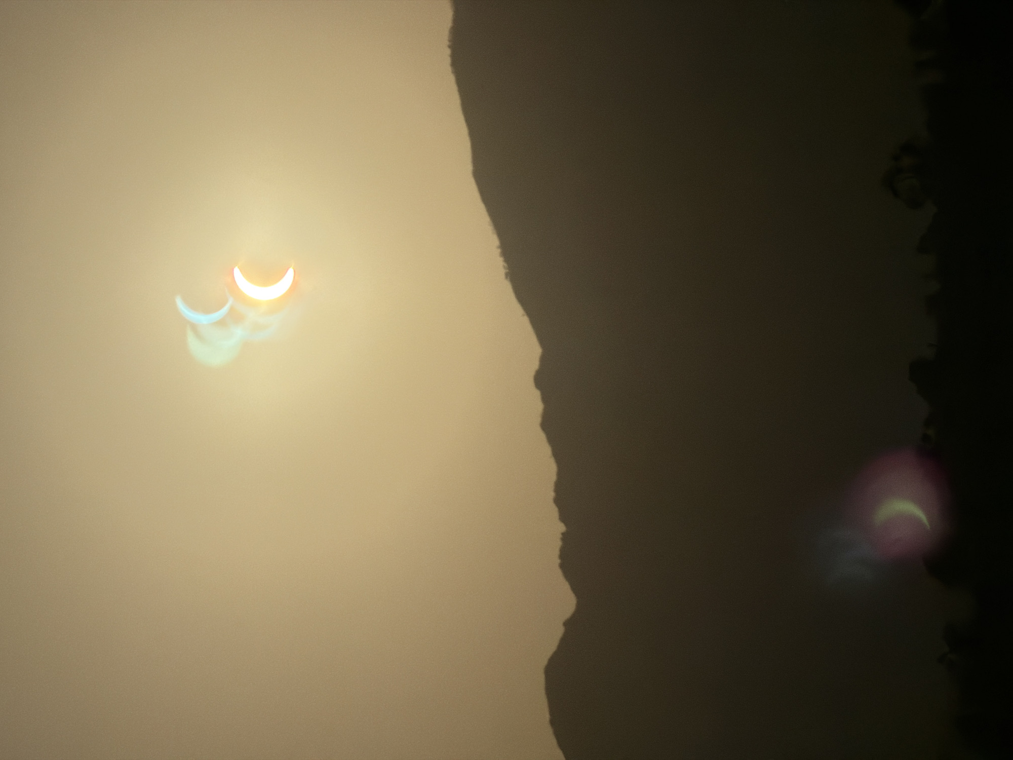 The image shows a silhouette of mountains against a hazy, golden sky. The sky is dominated by a solar eclipse, with multiple crescent-shaped images of the sun visible due to the effect of lens flare or multiple exposures. One prominent crescent is a bright orange and yellow, while others appear fainter and tinged with blue and green. In the foreground, a dark silhouette of trees and possibly low structures can be seen along the base of the mountains. The overall impression is that of a dramatic celestial event occurring over a natural landscape, potentially during sunset or sunrise given the color of the sky. There are no people visible in the image. The location of Resinera-voladilla, Spain, is mentioned as the setting, suggesting this is a view of the local mountains during the eclipse.