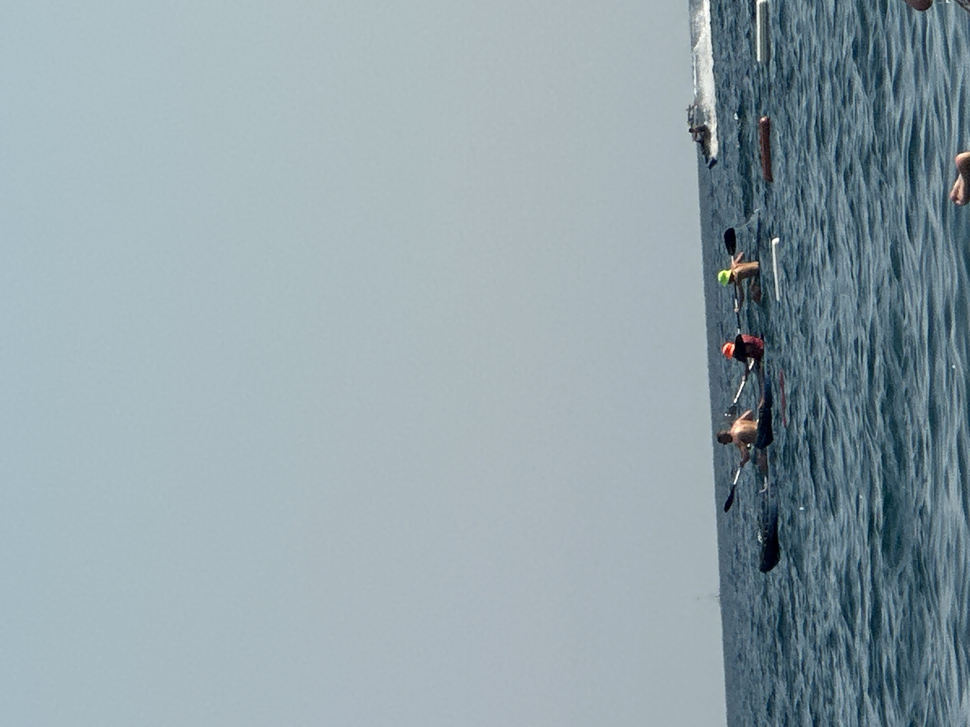 The image depicts a body of water, likely the sea, with several individuals engaged in water activities. In the foreground, there are at least three kayaks with people paddling. One kayak on the left has a person with their back to the camera, paddling with a double-bladed oar. To the right of this, another kayak is occupied by a person wearing an orange hat and a red shirt, also paddling. Further to the right, a third kayak has a person wearing a yellow hat and no shirt, paddling. Scattered in the water around the kayaks are some floating objects, including what appear to be white buoys or floats and a brown log-like object. In the background, a speedboat is visible, creating a wake as it moves across the water. The sky is clear and pale blue. The overall scene suggests a recreational day on the water. The location is identified as the city of Boreti, Montenegro.